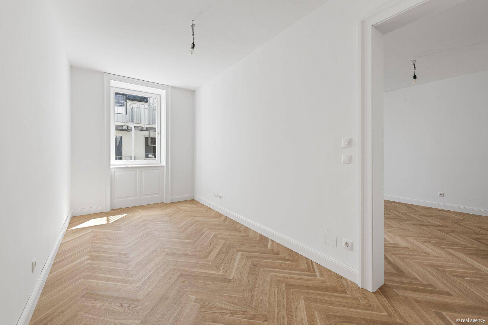 Ruhiger Raum mit Fischgrätparkett und einem großen Fenster, ideal als Schlaf- oder Arbeitszimmer.