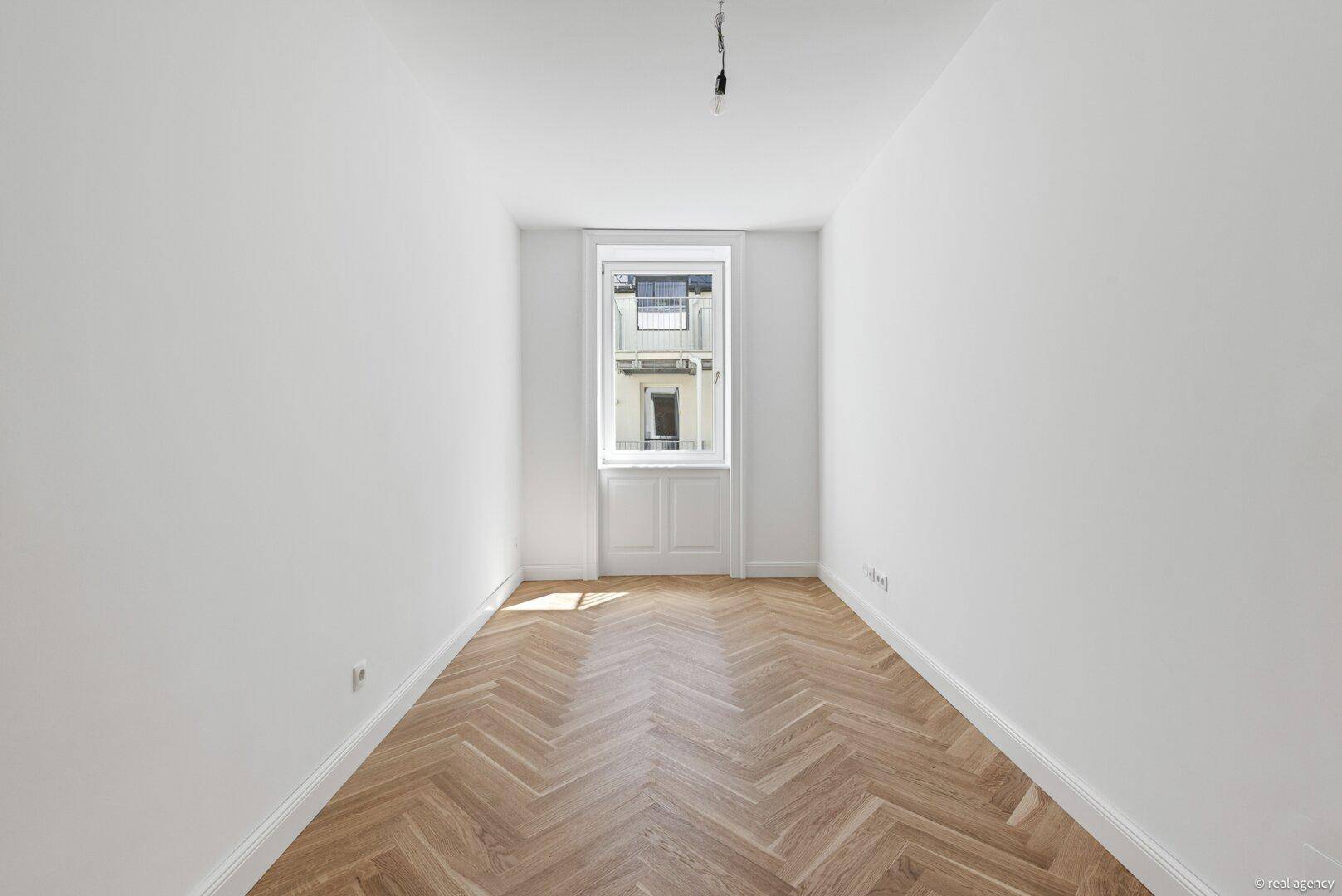 Schmaler, heller Raum mit Fischgrätparkett und einem Fenster, geeignet als Schlafzimmer oder Büro.