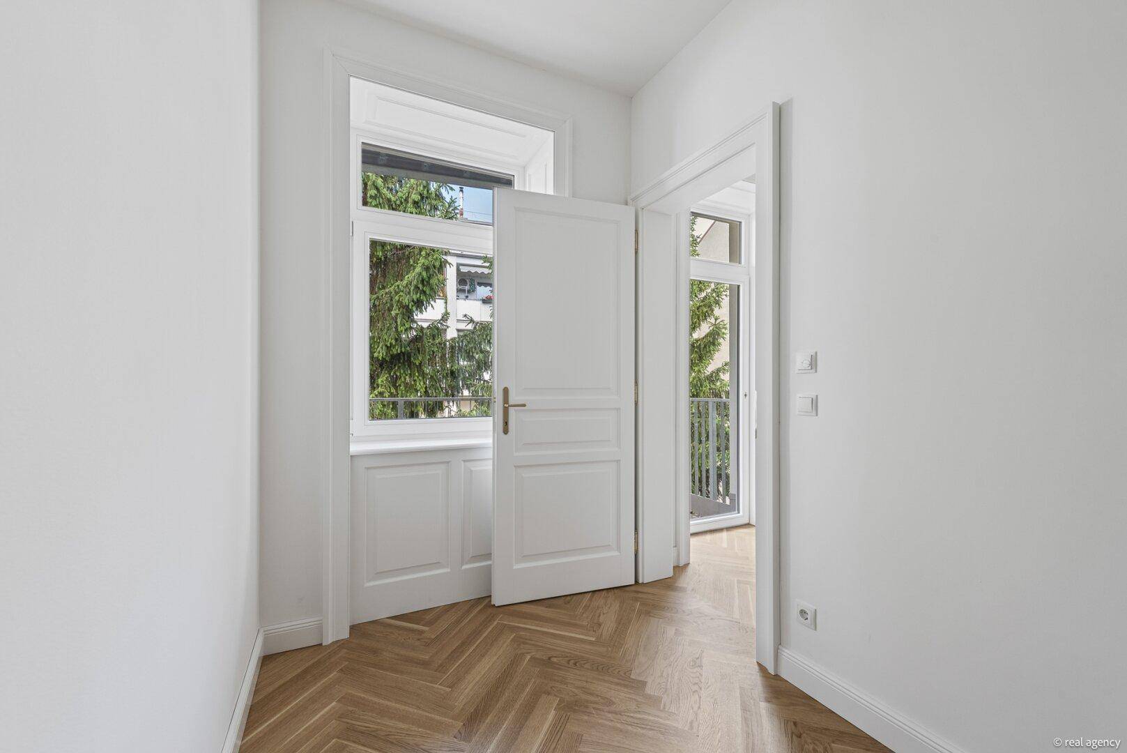 Helles Zimmer mit Fischgrätparkett, hohen Fenstern und Zugang zu einem sonnigen Balkon.