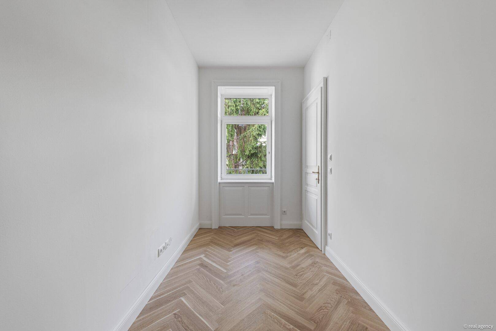 Schmaler Raum mit Fischgrätparkett und einem hohen Fenster, ideal als Schlaf- oder Arbeitszimmer.