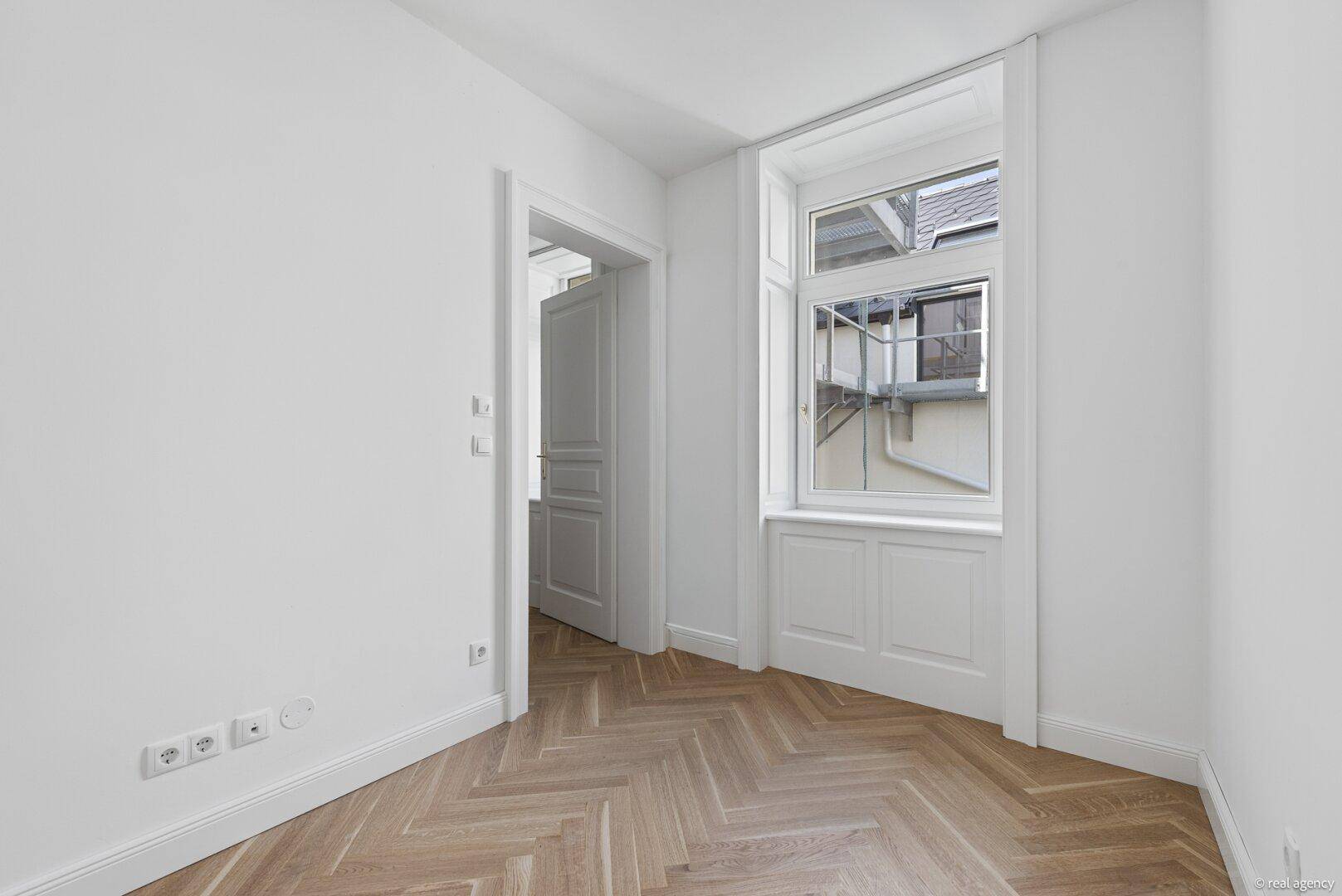 Leerer Raum mit Fischgrätparkettboden und einem Fenster mit Blick auf den Innenhof.