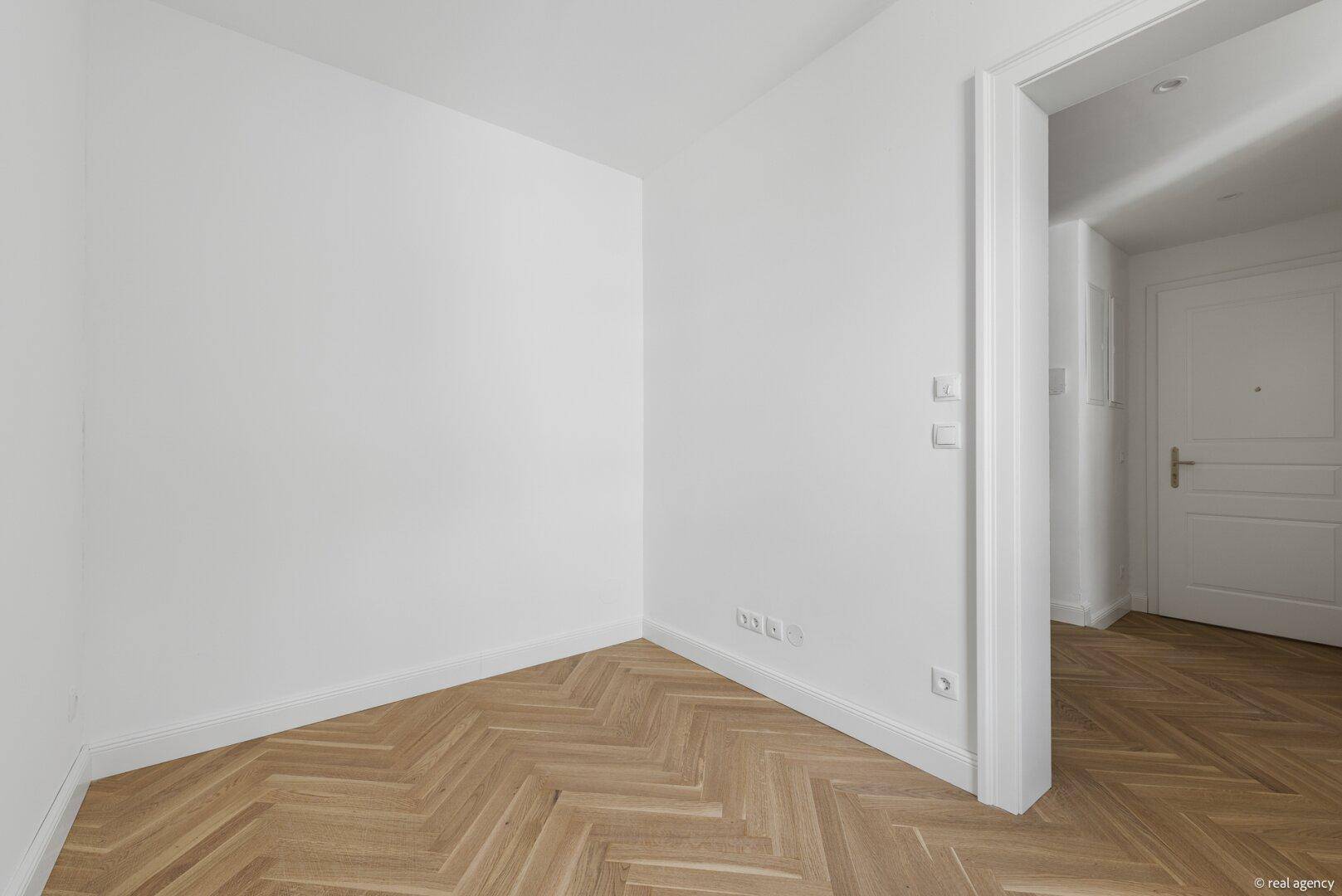Heller, leerer Raum mit Fischgrätparkettboden und Zugang zu einem weiteren Zimmer.