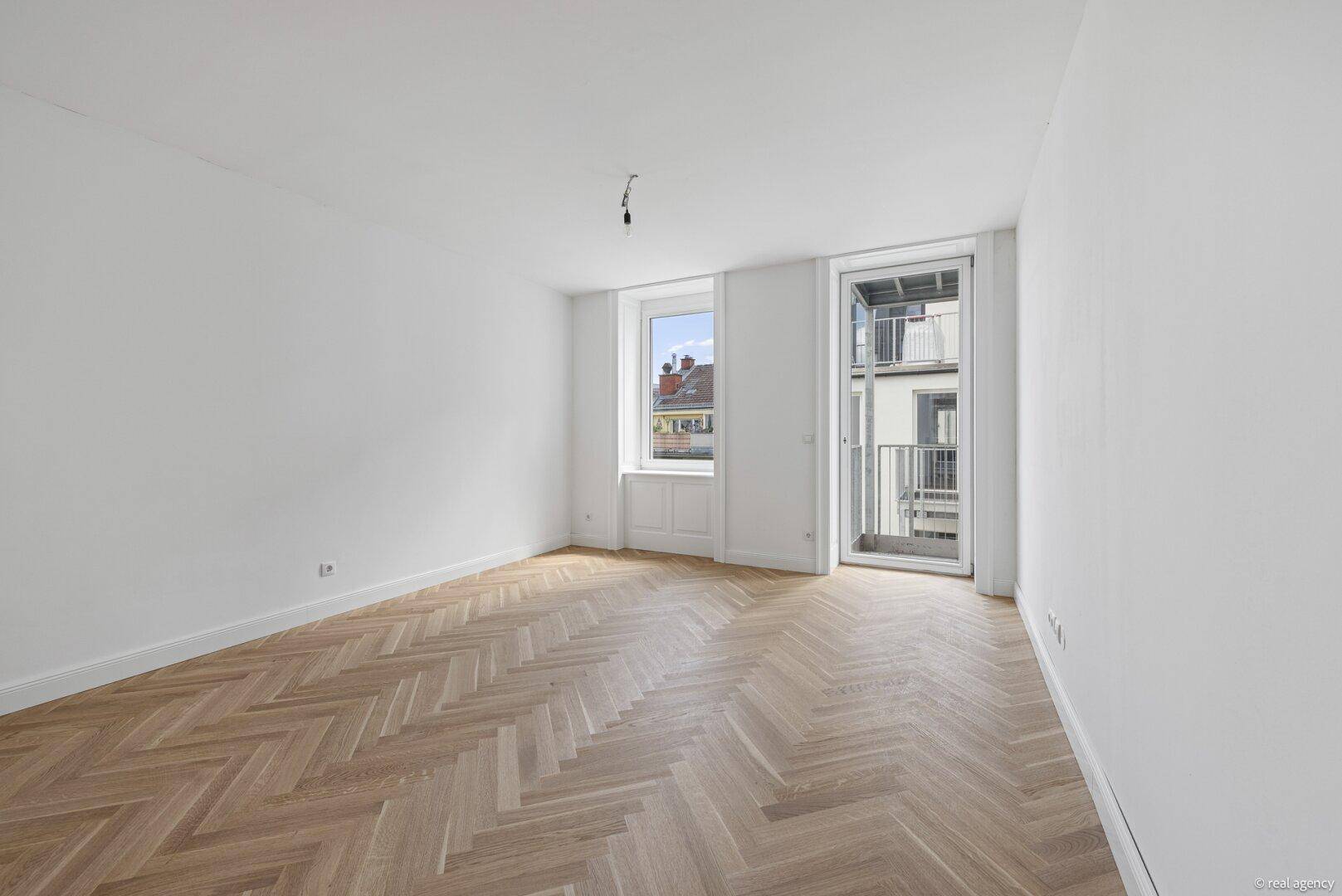 Geräumiger, heller Raum mit Fischgrätparkett und Zugang zu einem kleinen Balkon.