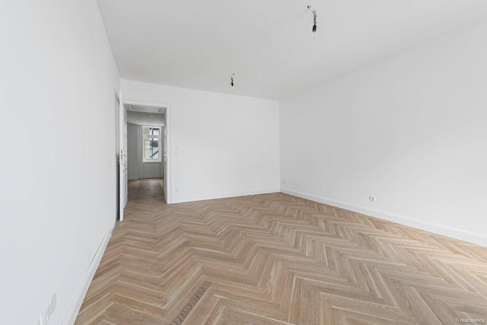 Heller, leerer Raum mit Fischgrätparkettboden und einem Durchgang zu einem weiteren Zimmer.