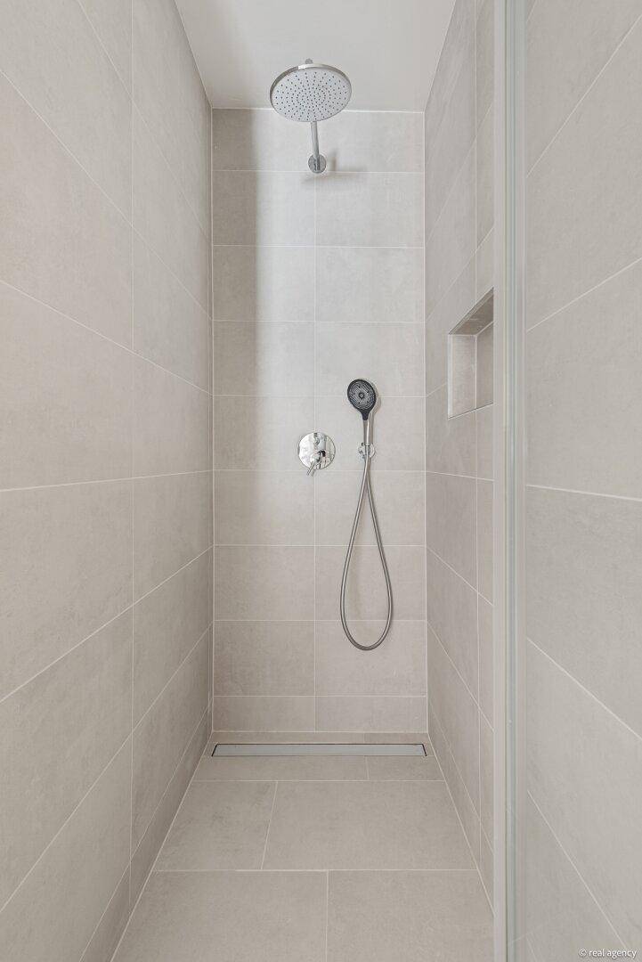 Geräumige begehbare Dusche mit Regenduschkopf und Handbrause, komplett gefliest in Grau.