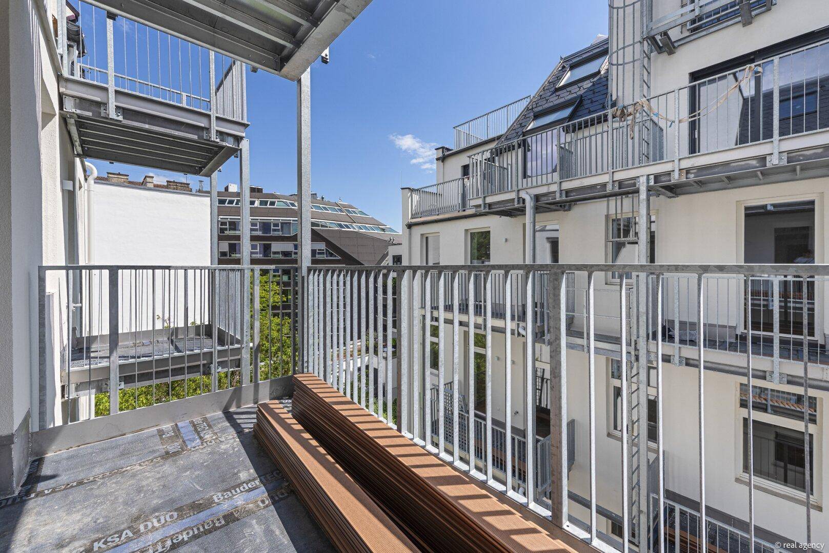Heller Balkon mit Metallgeländer und freiem Blick auf die Nachbargebäude unter blauem Himmel.