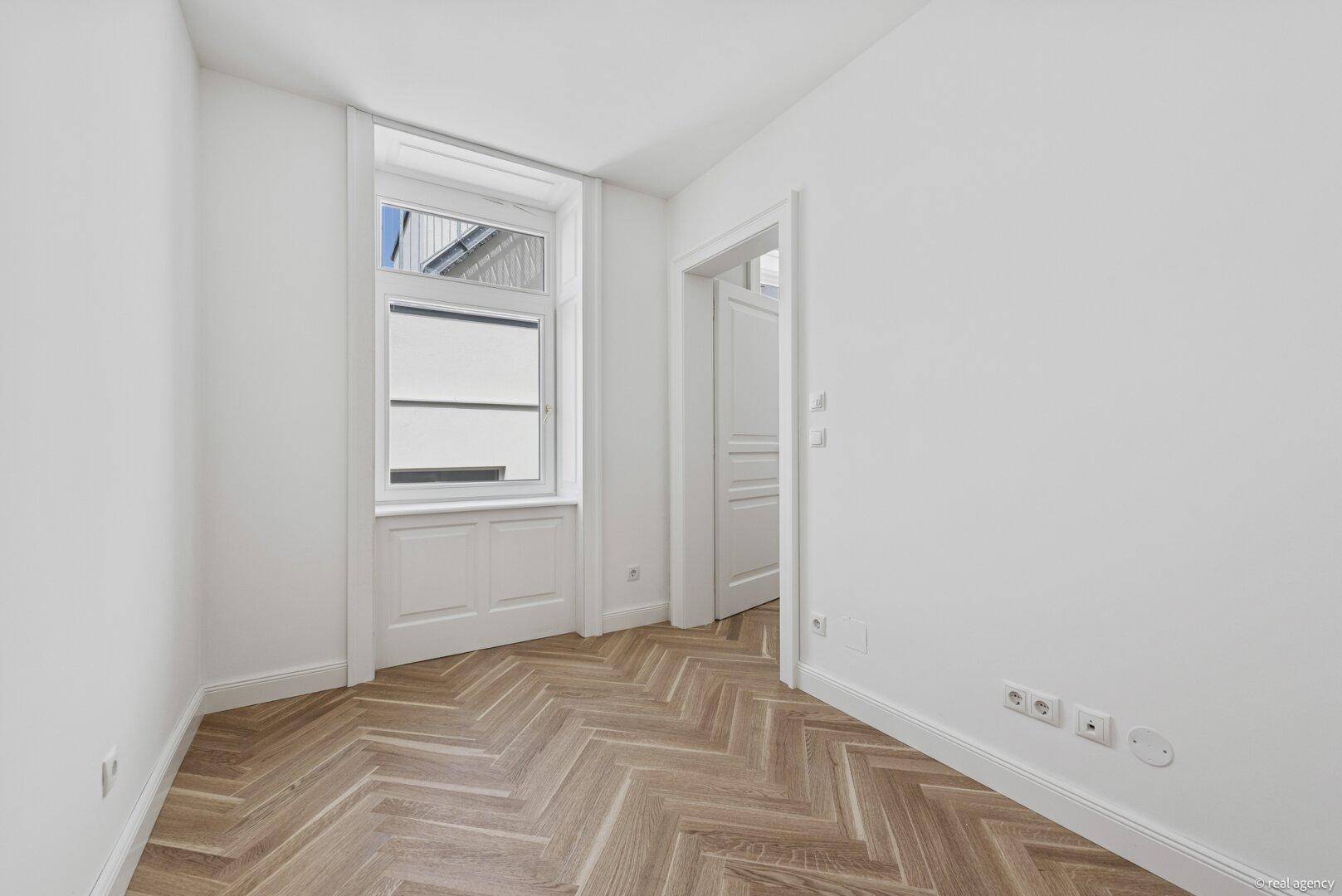Helles Zimmer mit Fischgrätparkett und einem hohen Fenster, ideal als Schlaf- oder Arbeitszimmer.