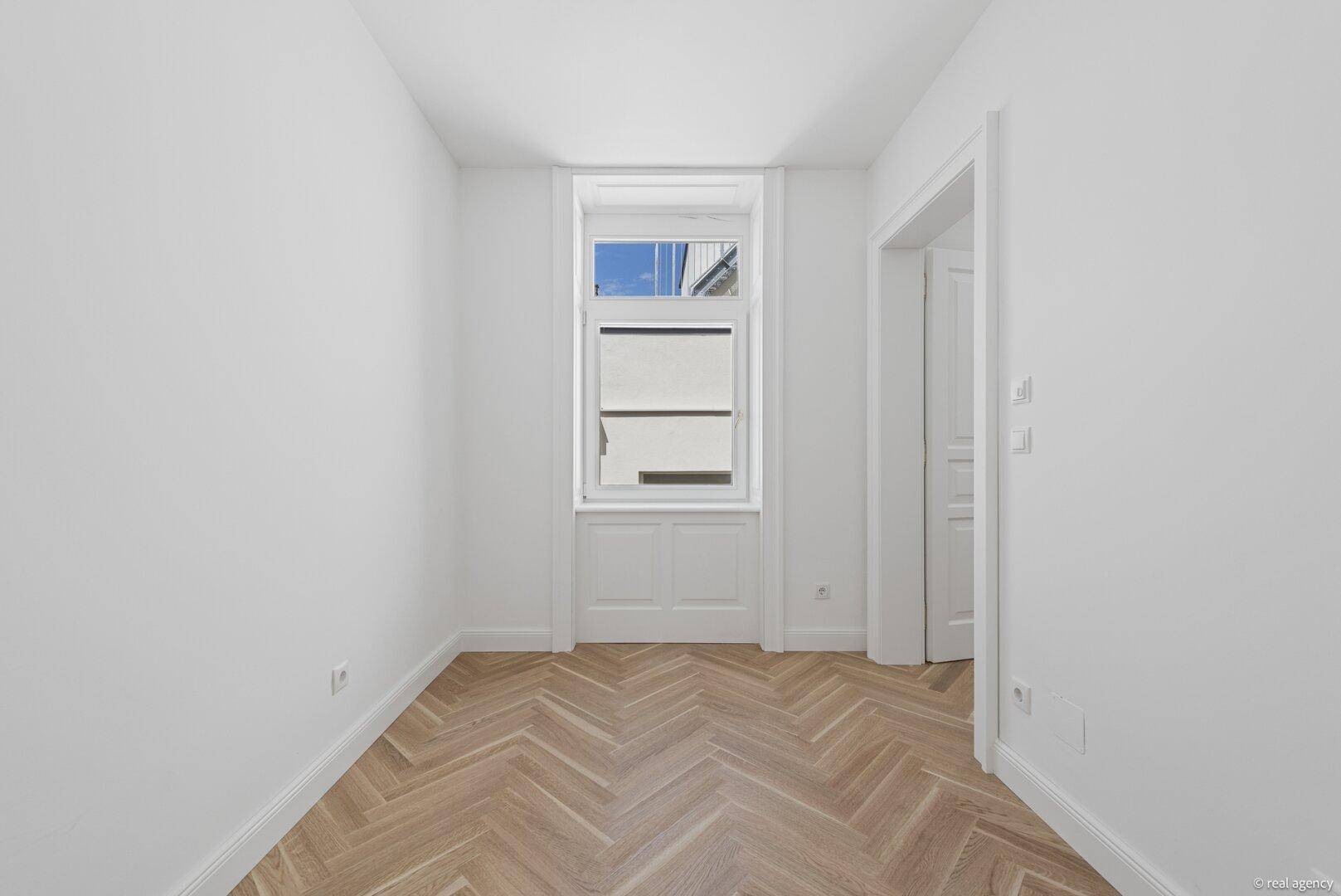 Leeres, helles Zimmer mit Fischgrätparkett und weiß gestrichenen Wänden, mit Blick nach draußen.