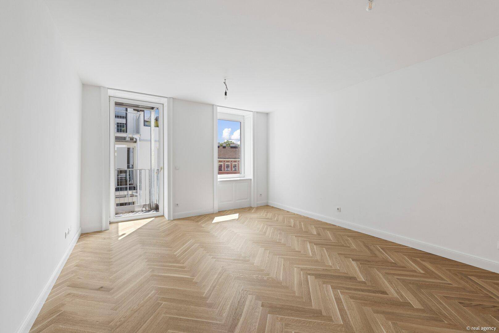 Helles, leeres Zimmer mit Fischgrätparkett und Zugang zu einem französischen Balkon.