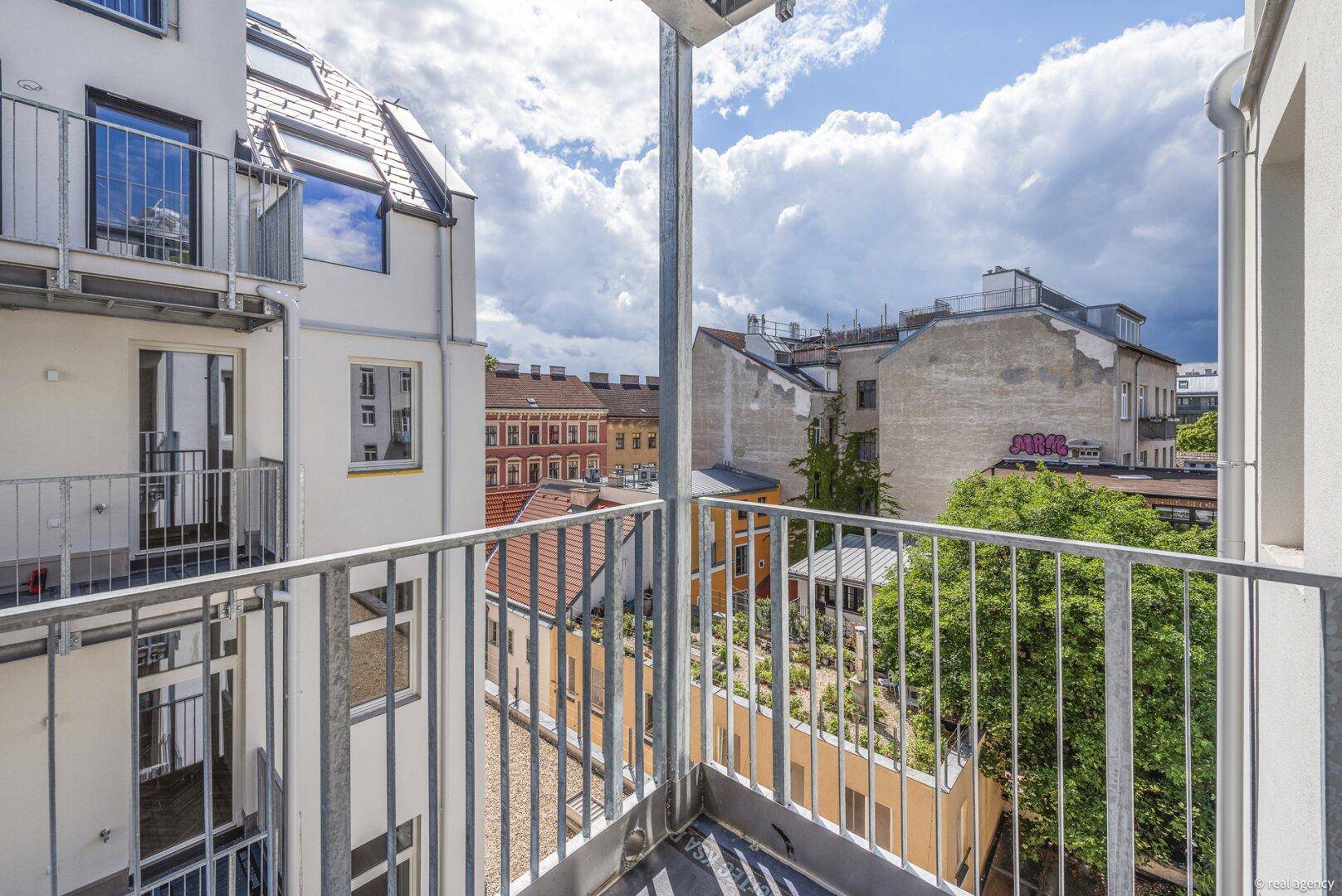 Blick vom Balkon über die Dächer der Stadt mit grünen Bäumen und bewölktem Himmel.