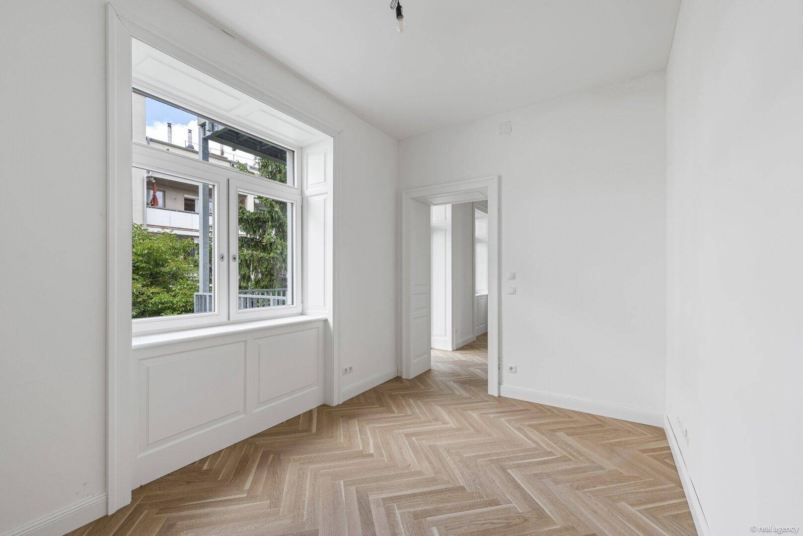 Helles, leeres Zimmer mit Fischgrätparkett und Blick ins Grüne.