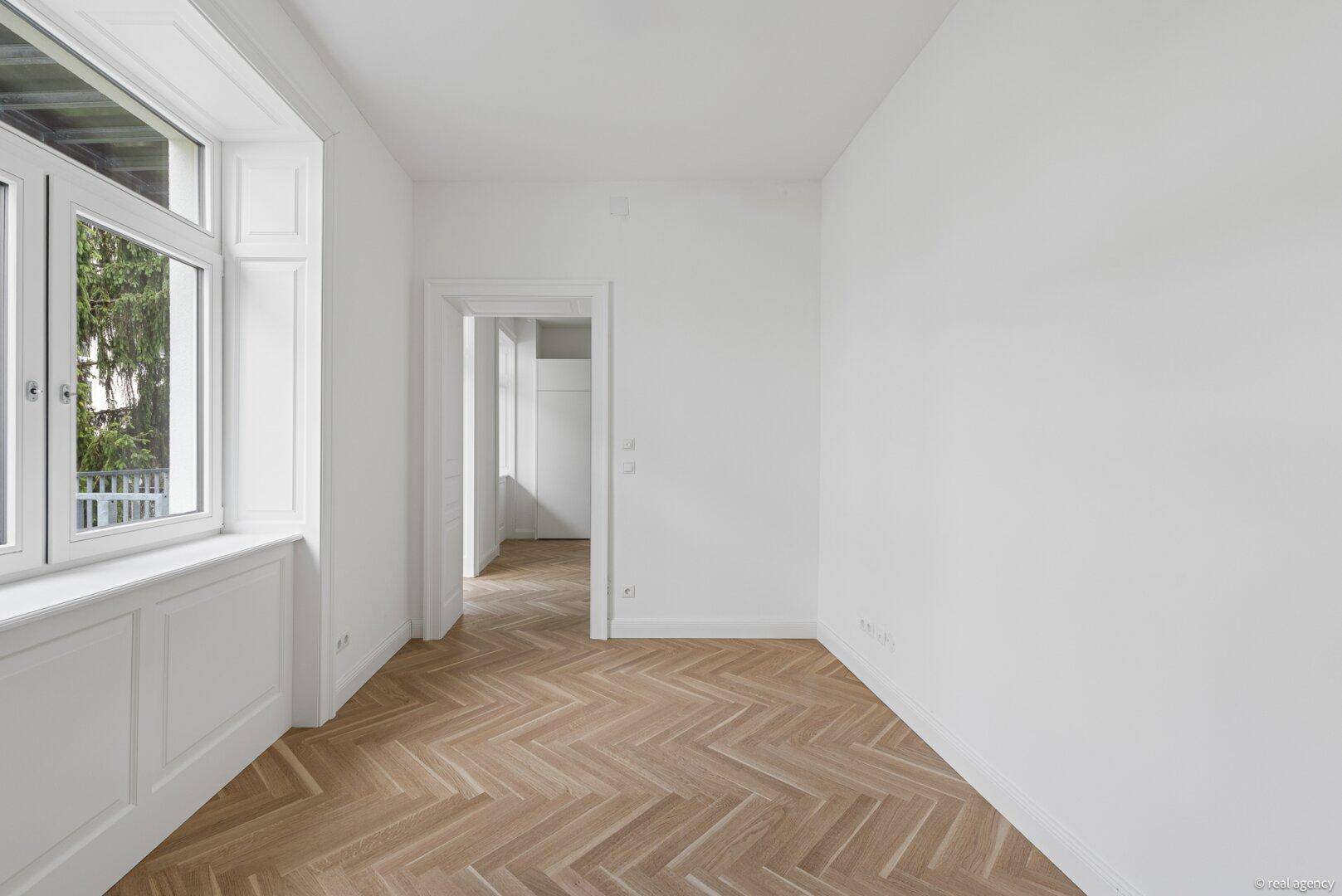 Geräumiges, leeres Zimmer mit Fischgrätparkett und weißen Wänden.