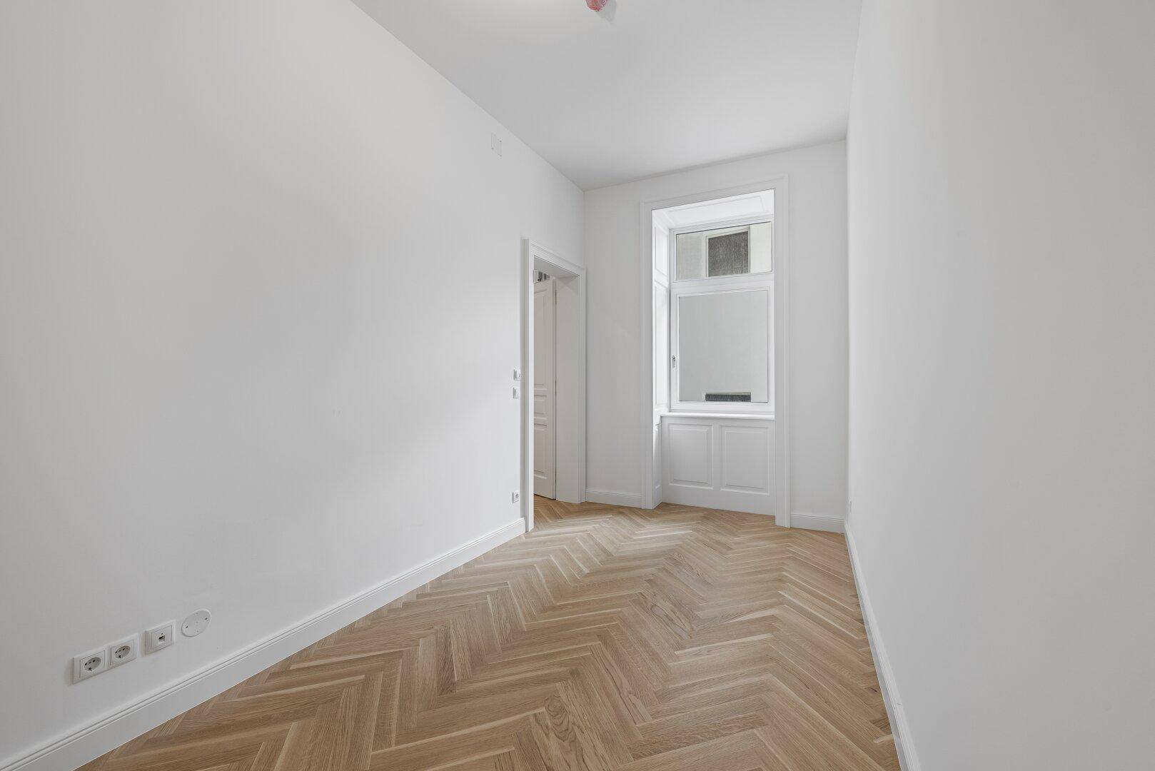 Leerer Raum mit hellem Fischgrätparkettboden, weißen Wänden und einem klassischen Fenster.