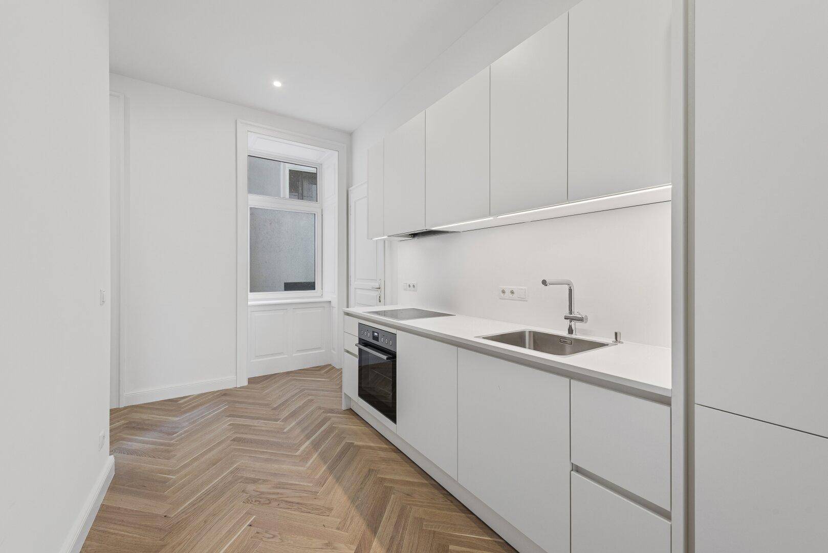 Moderne, helle Küchenzeile mit weißen Schränken, integriertem Backofen und Fischgrätparkettboden.
