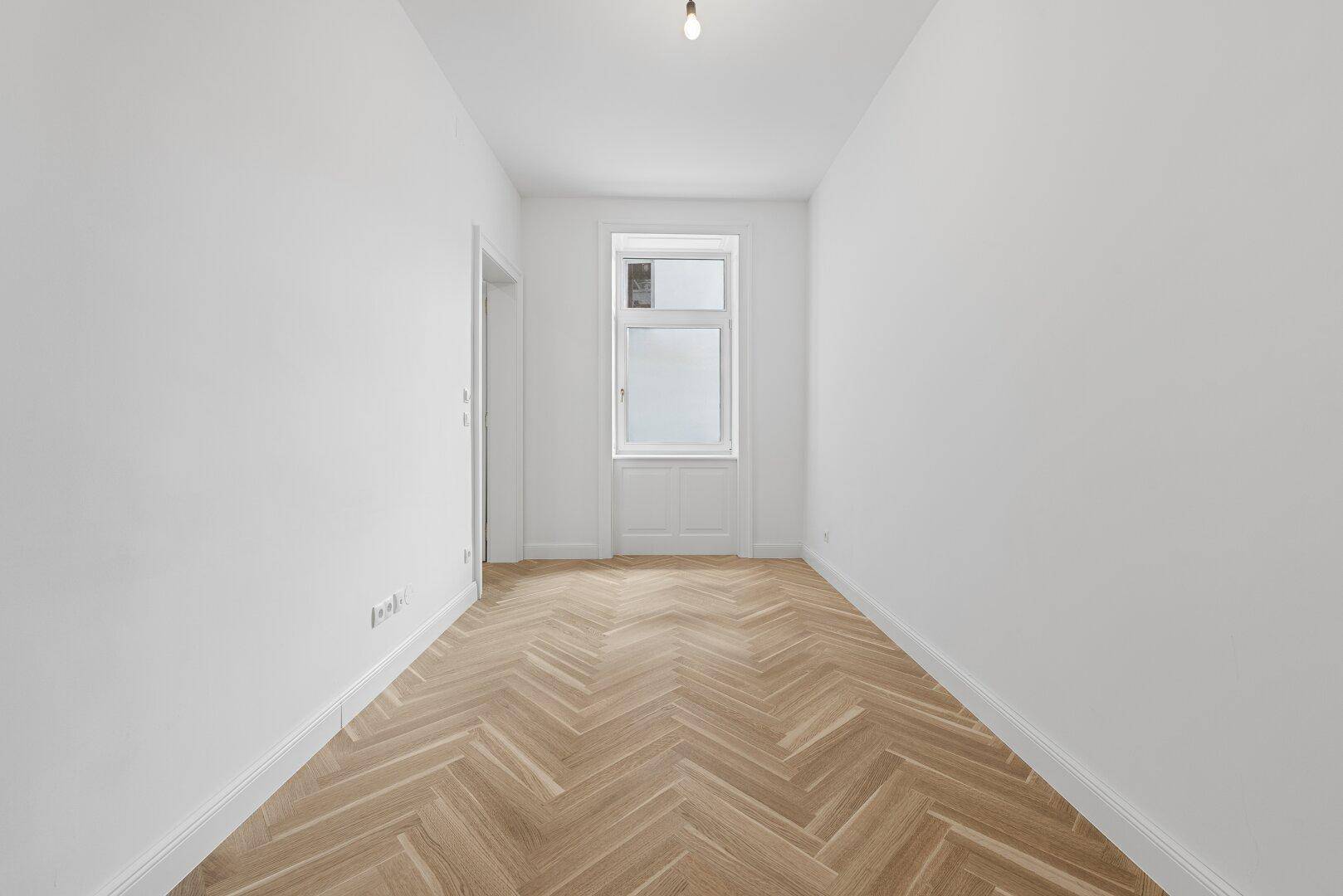Schmaler, heller Raum mit Fischgrätparkett und einem hohen Fenster, bietet viel Tageslicht.