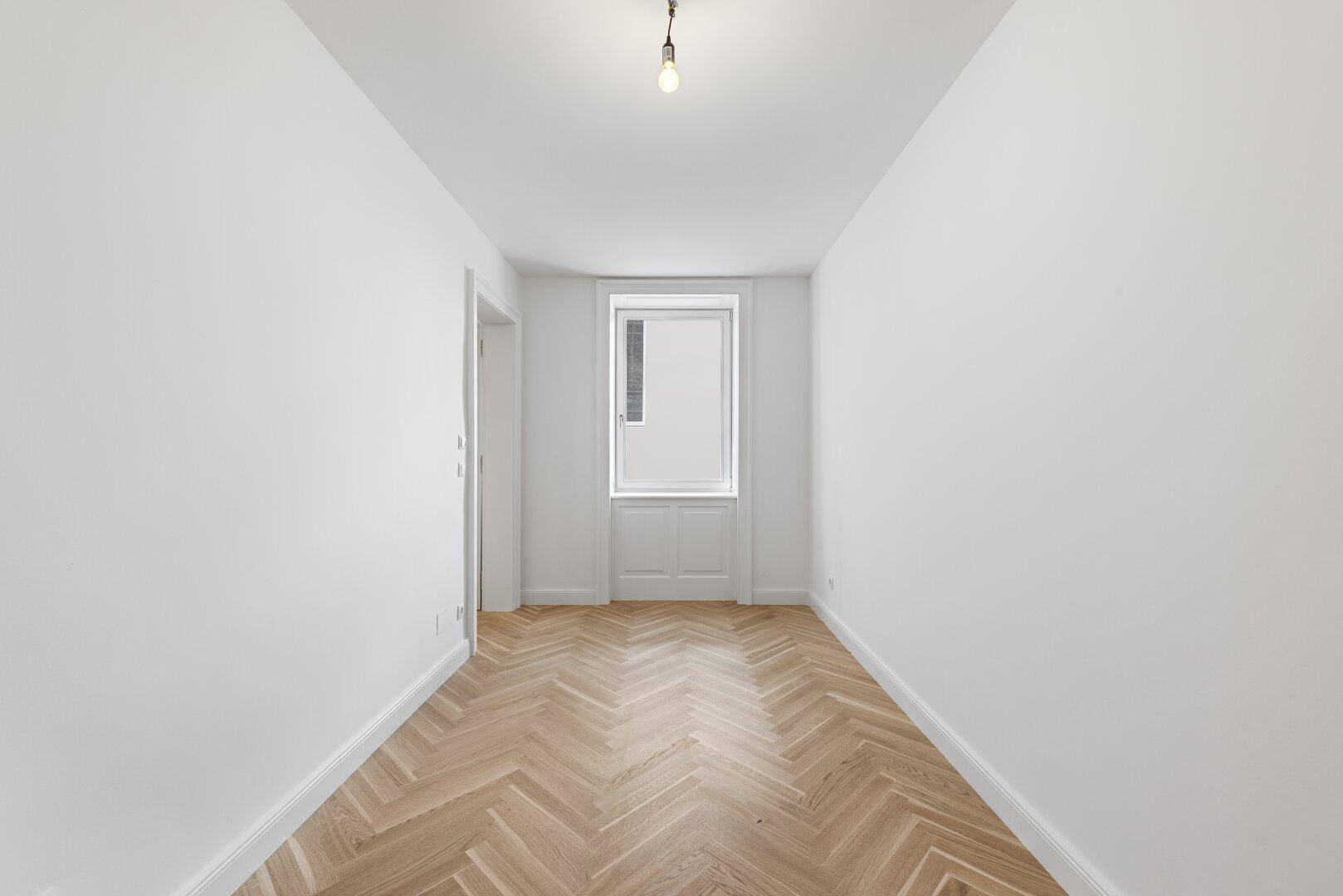 Leeres, helles Zimmer mit Fischgrätparkett und einem Fenster, das viel Tageslicht hereinlässt.