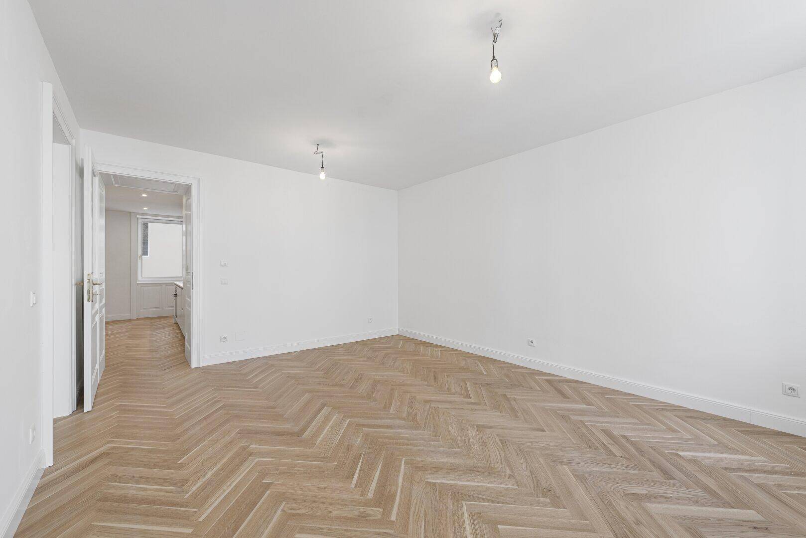 Geräumiger, heller Raum mit Fischgrätparkett und Blick in die angrenzende Küche.