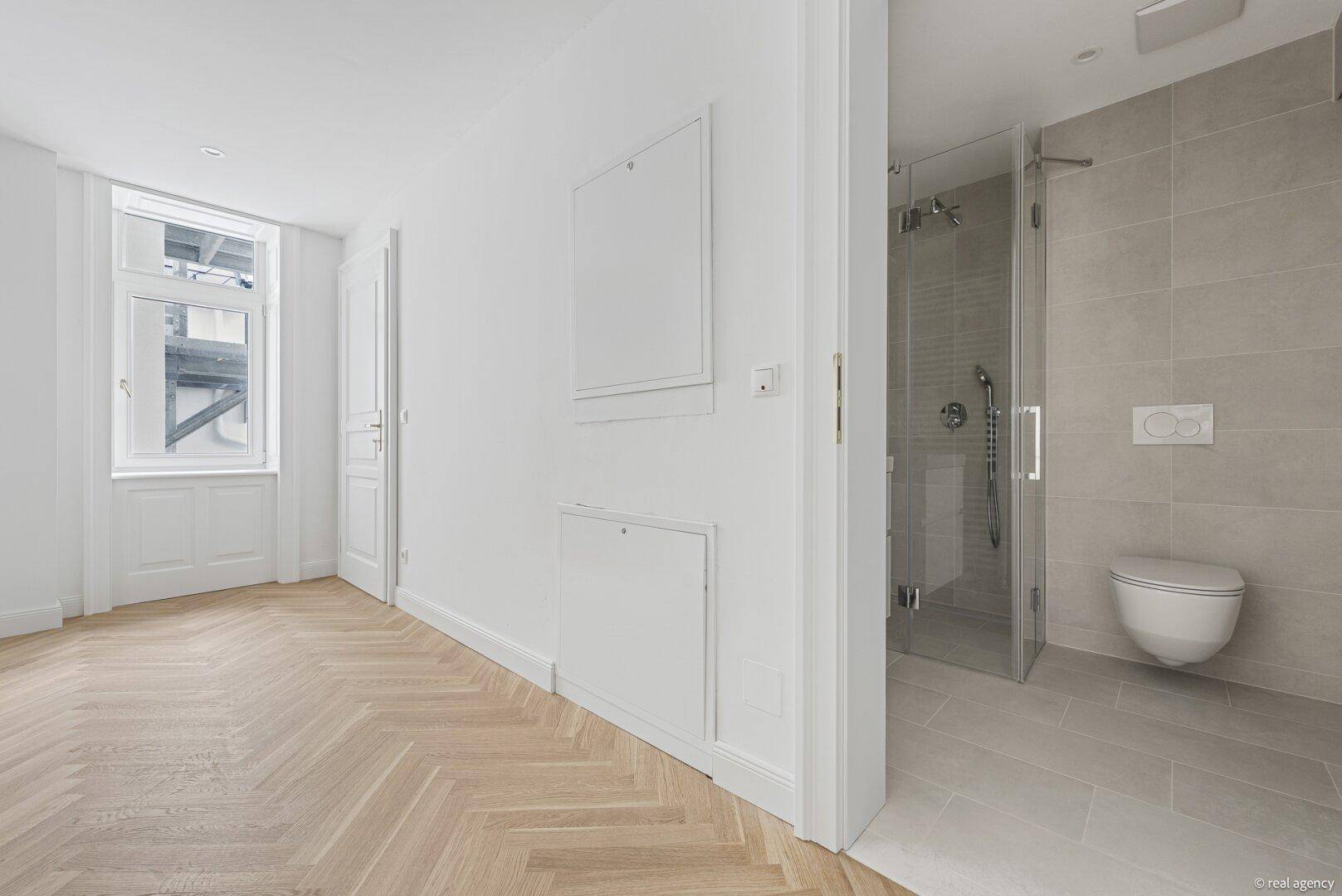 Heller Raum mit Fischgrätparkett und direktem Zugang zu einem modernen Badezimmer mit Dusche.