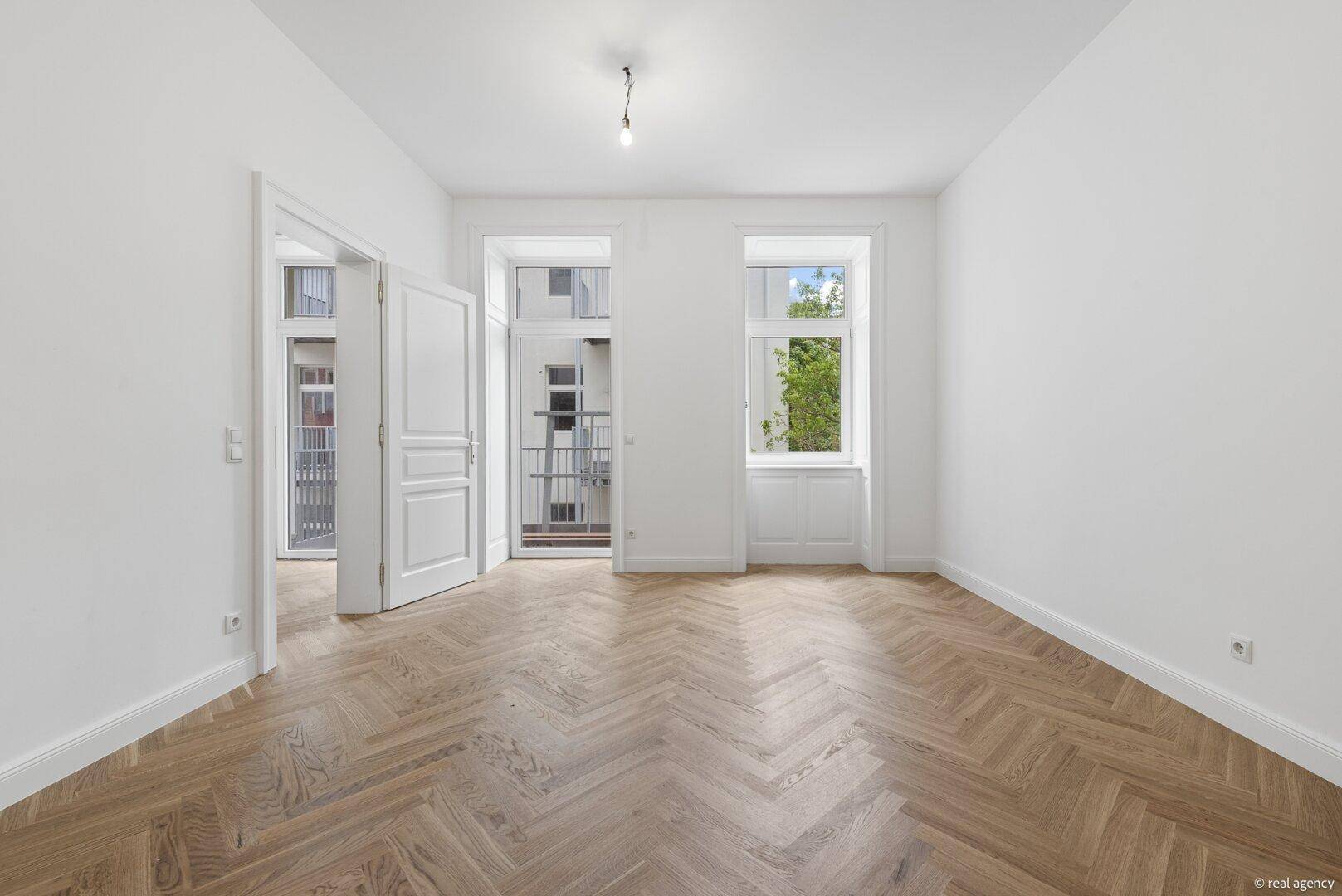 Heller Raum mit Fischgrätparkett, weißen Wänden und Zugang zu einem französischen Balkon.