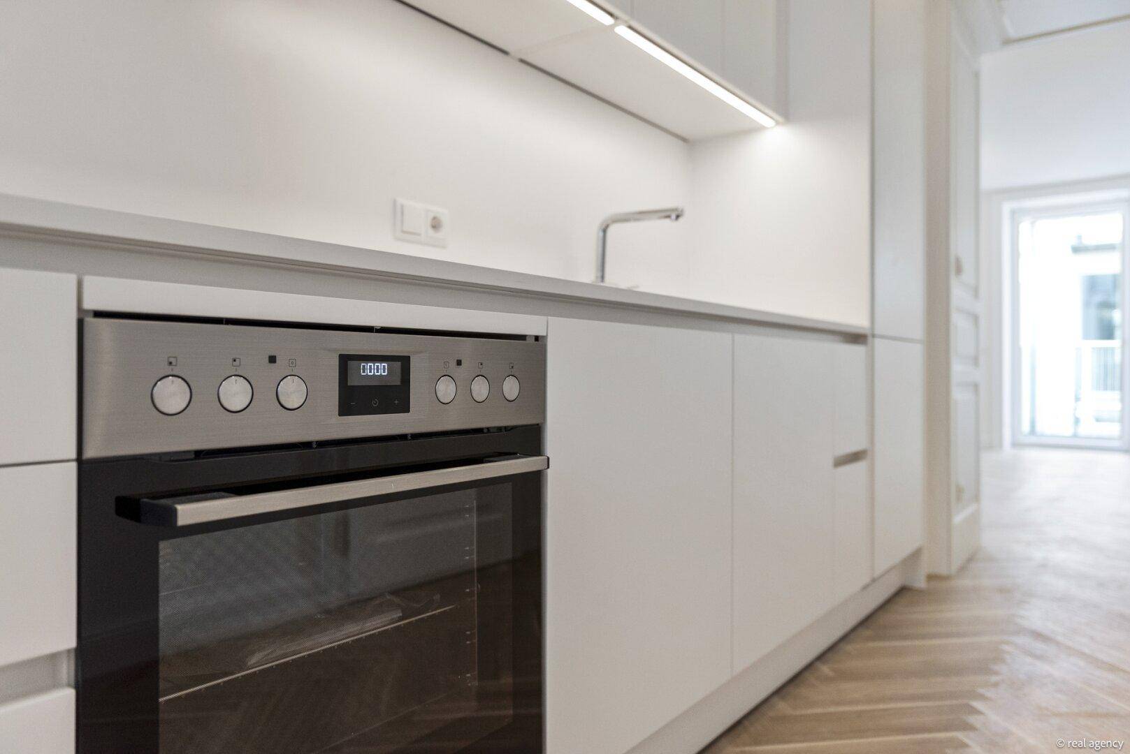 Moderne Einbauküche mit integriertem Backofen und weißen Schränken, daneben Fischgrätparkett.