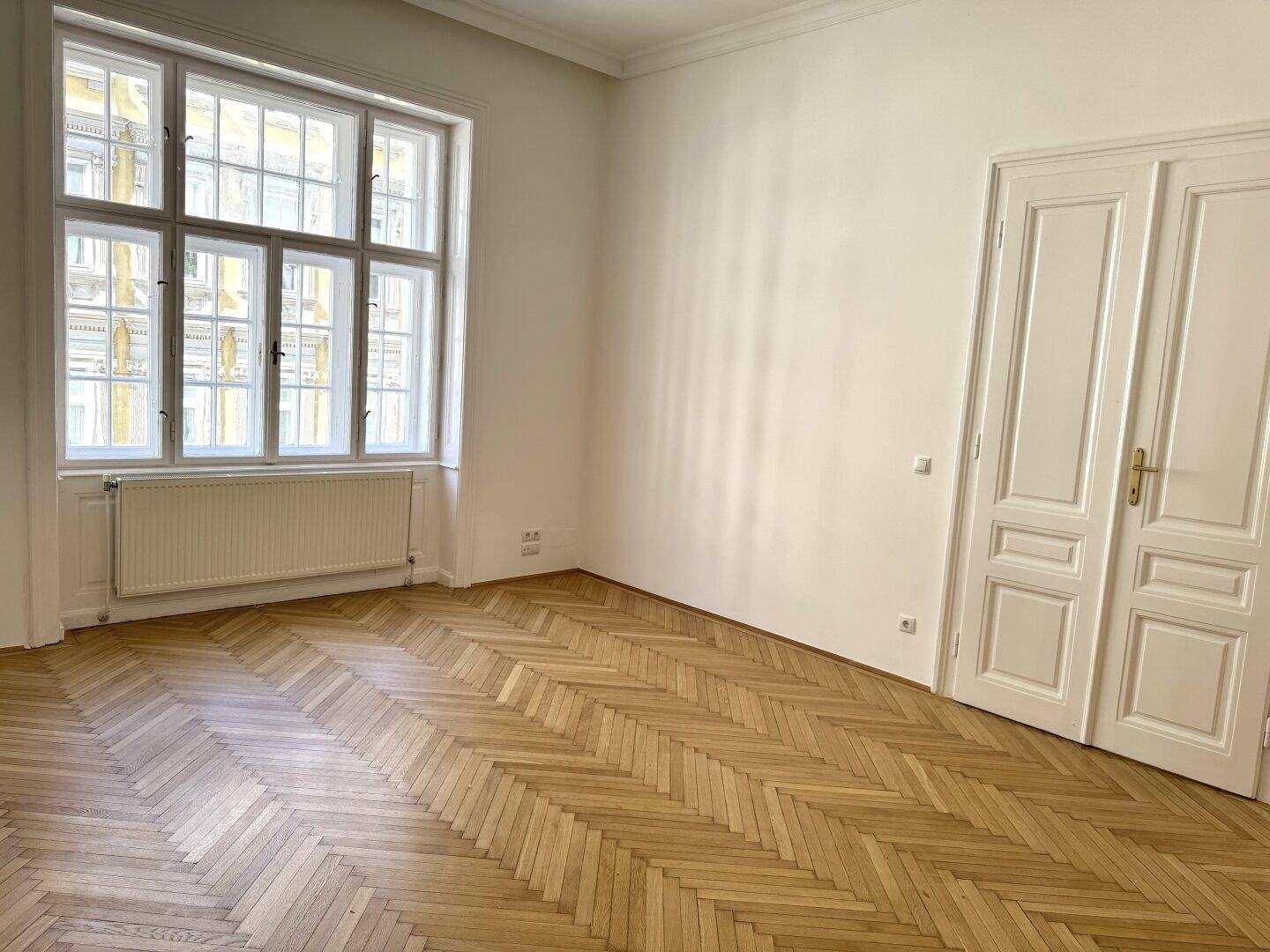 Großzügiges Zimmer mit Fischgrätparkett, hohen Decken und einem großen Fenster, das viel Licht spendet.