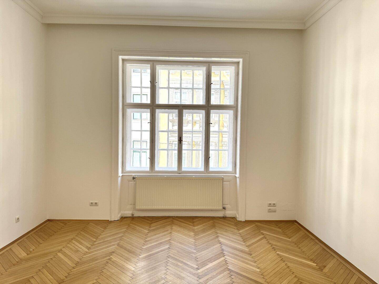 Geräumiges Zimmer mit Fischgrätparkett, hohen Decken und einem großen Fenster, das den Raum hell erscheinen lässt.