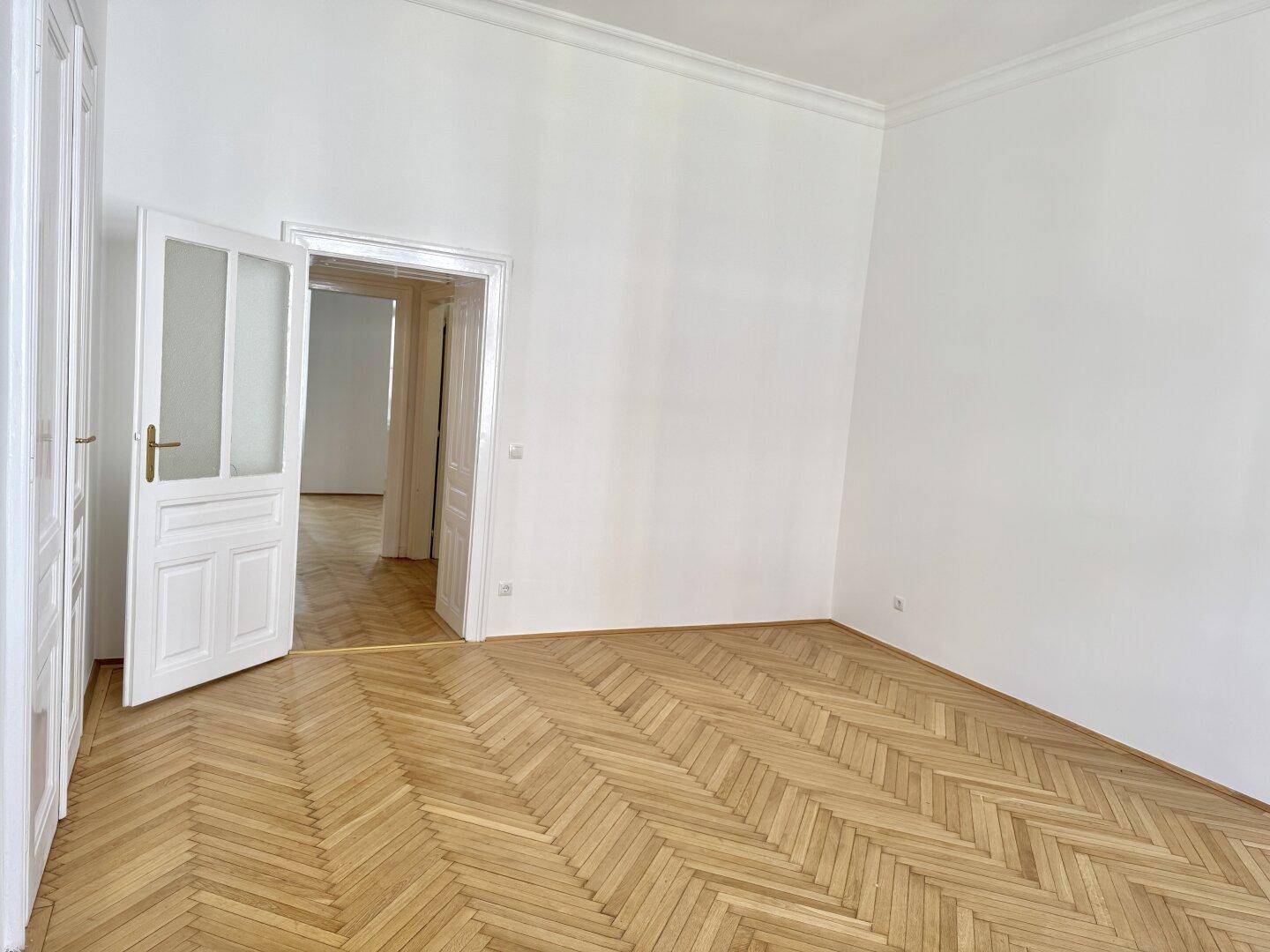 Helles Zimmer mit Fischgrätparkett und hohen Decken, das durch eine Glastür zu einem weiteren Raum führt.