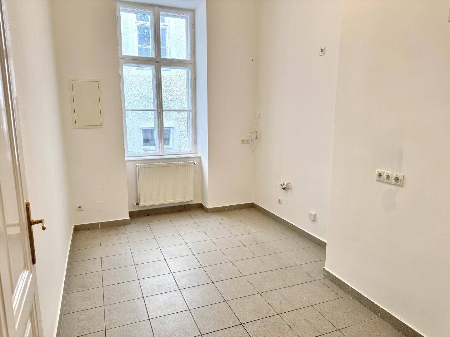 Heller Raum mit Fliesenboden und Fenster, vorbereitet für die Installation einer Küche mit sichtbaren Anschlüssen.