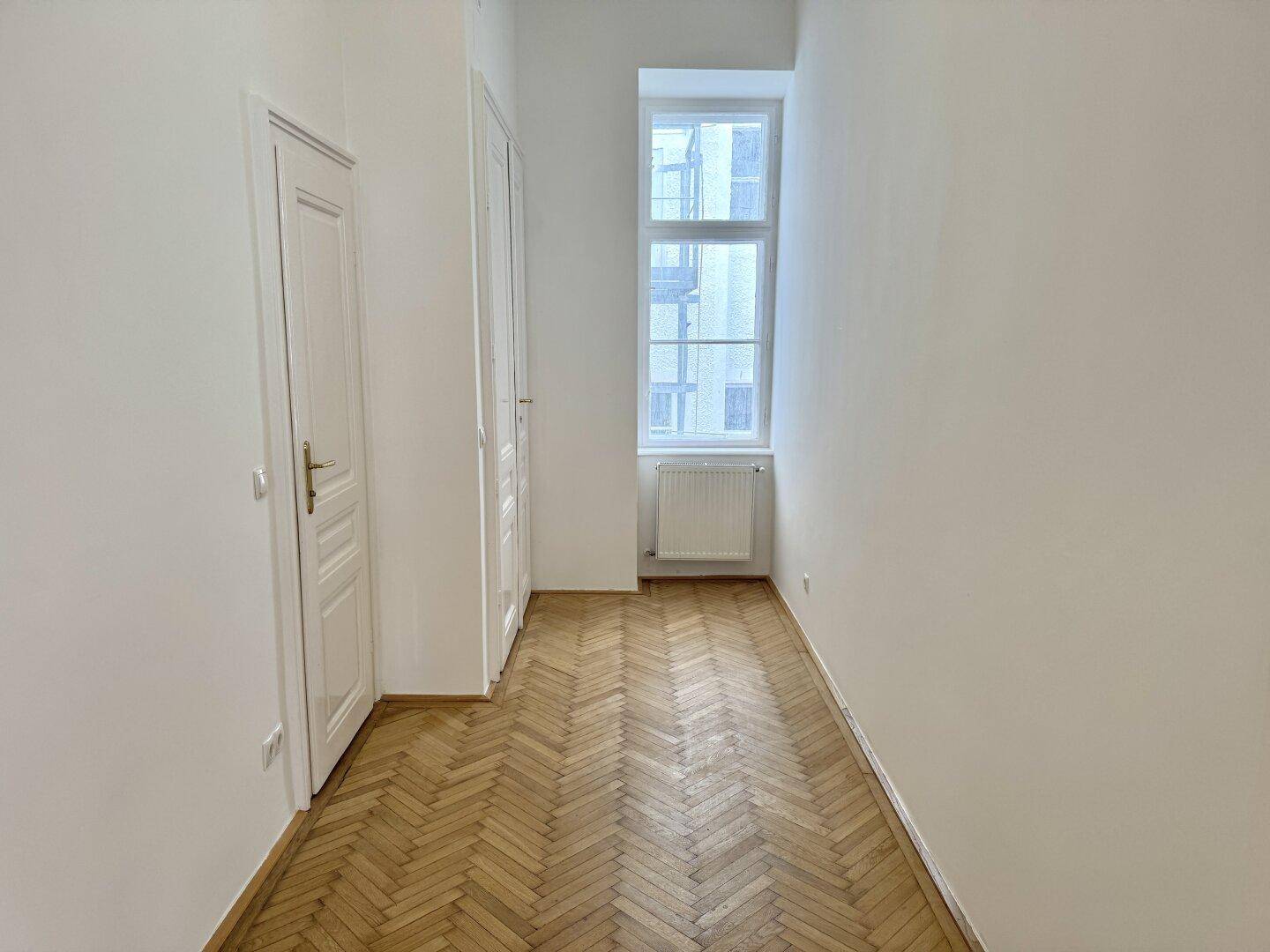 Schmaler Raum mit Fischgrätparkett, hohen Decken und einem Fenster, geeignet als Schlaf- oder Arbeitszimmer.
