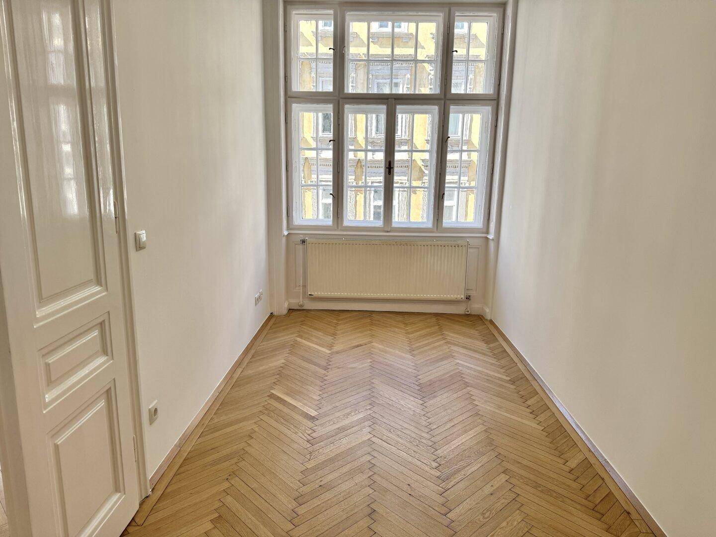 Helles Zimmer mit Fischgrätparkett und einem großen Fenster, das sich gut als Schlaf- oder Arbeitszimmer eignet.