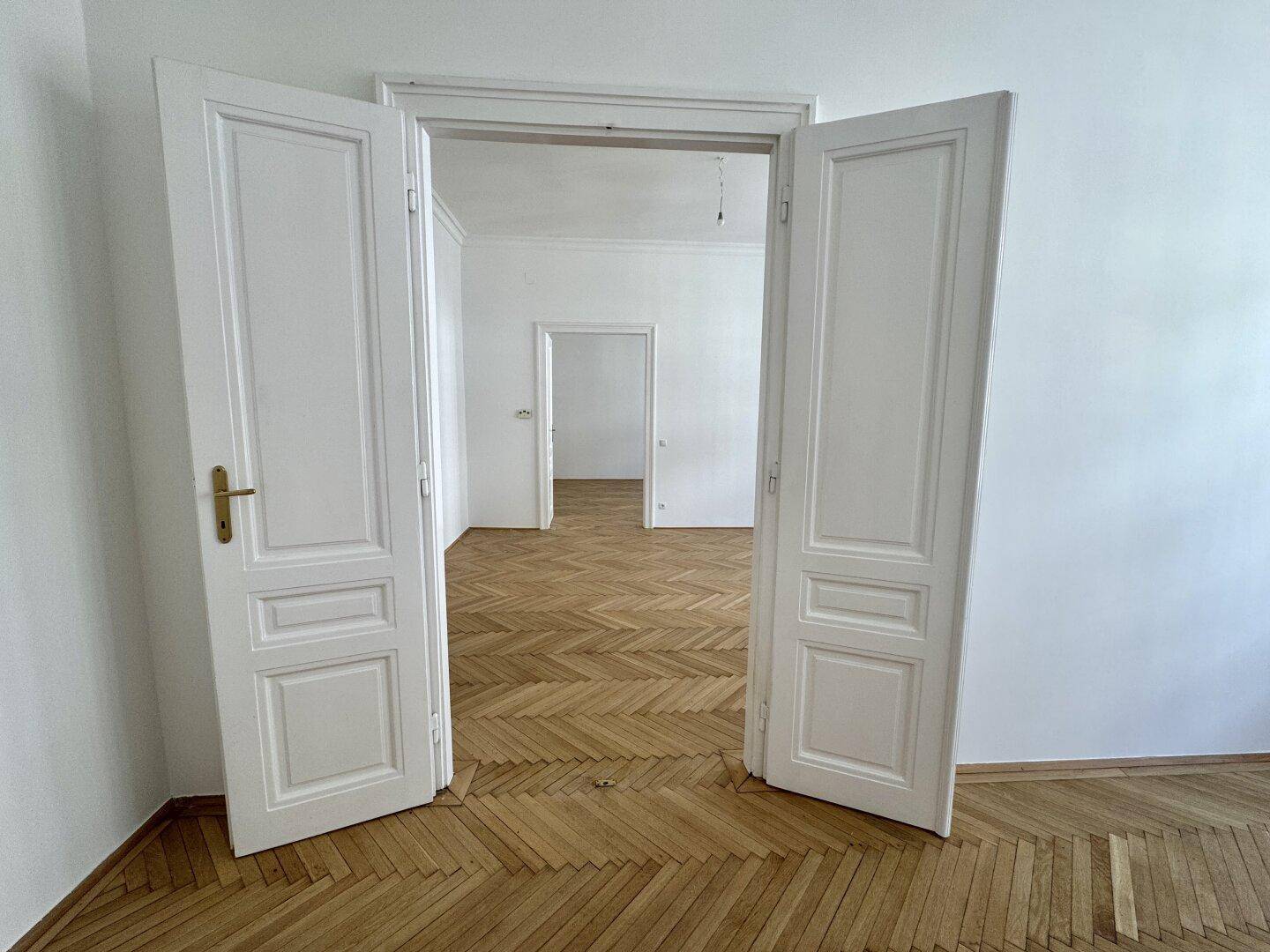 Helles Zimmer mit Fischgrätparkett und hohen Decken, das durch eine weit geöffnete Doppeltür weitere Räume zeigt.