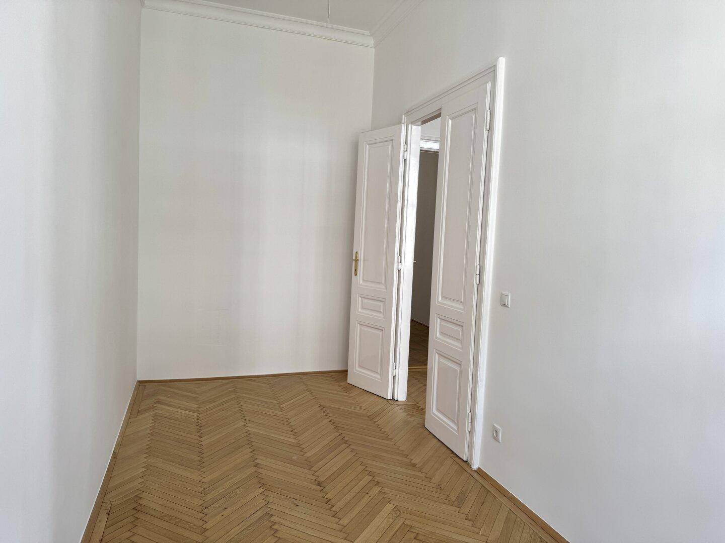 Geräumiges Zimmer mit Fischgrätparkett und hohen Decken, das durch eine Doppeltür zugänglich ist.