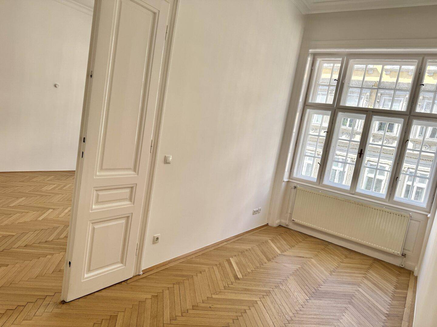 Heller Raum mit Fischgrätparkett und einem großen Fenster, das viel Tageslicht in den Wohnbereich bringt.