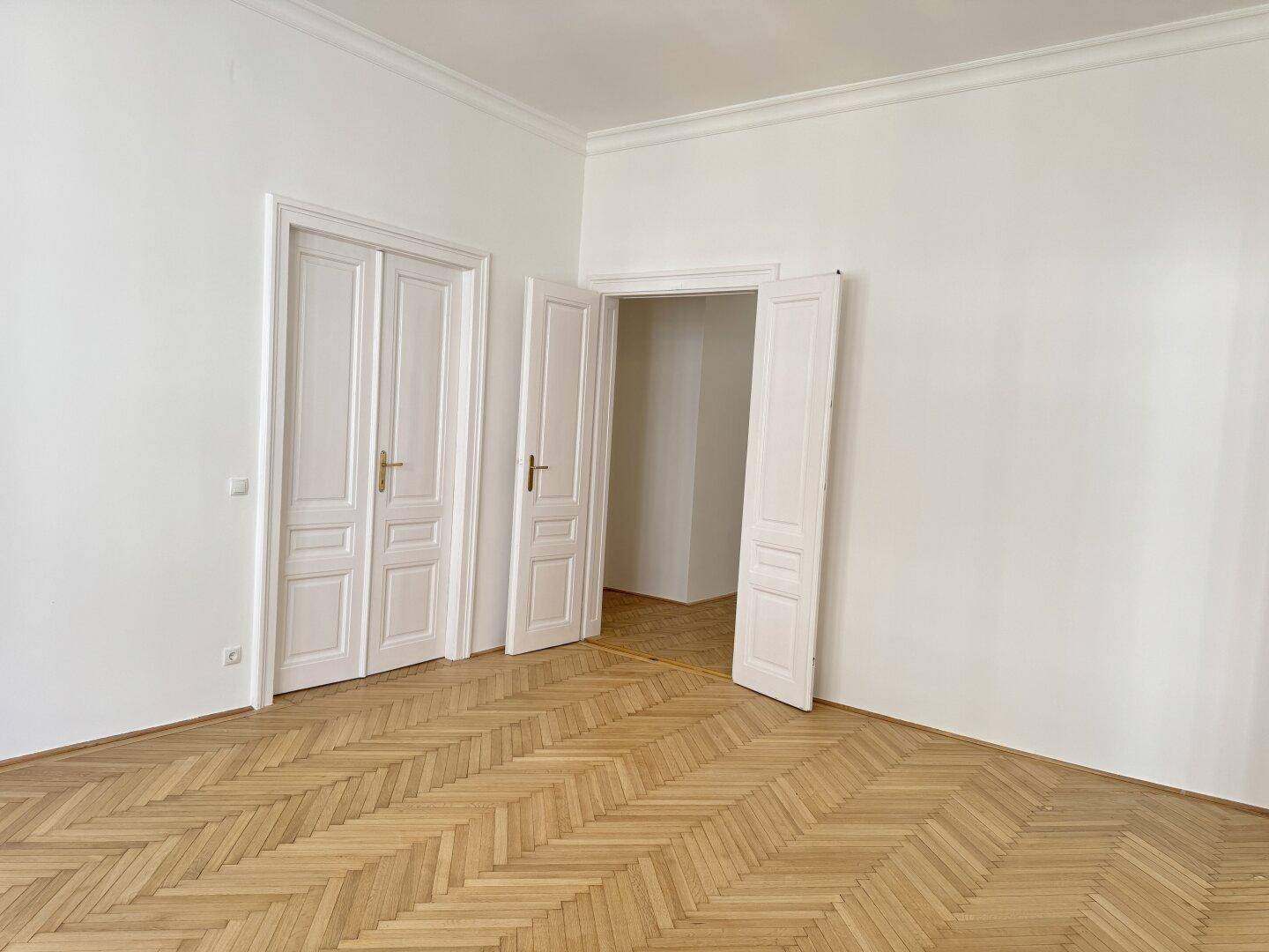 Helles Zimmer mit Fischgrätparkett und hohen Decken, verbunden durch eine Doppeltür zu einem weiteren Raum.