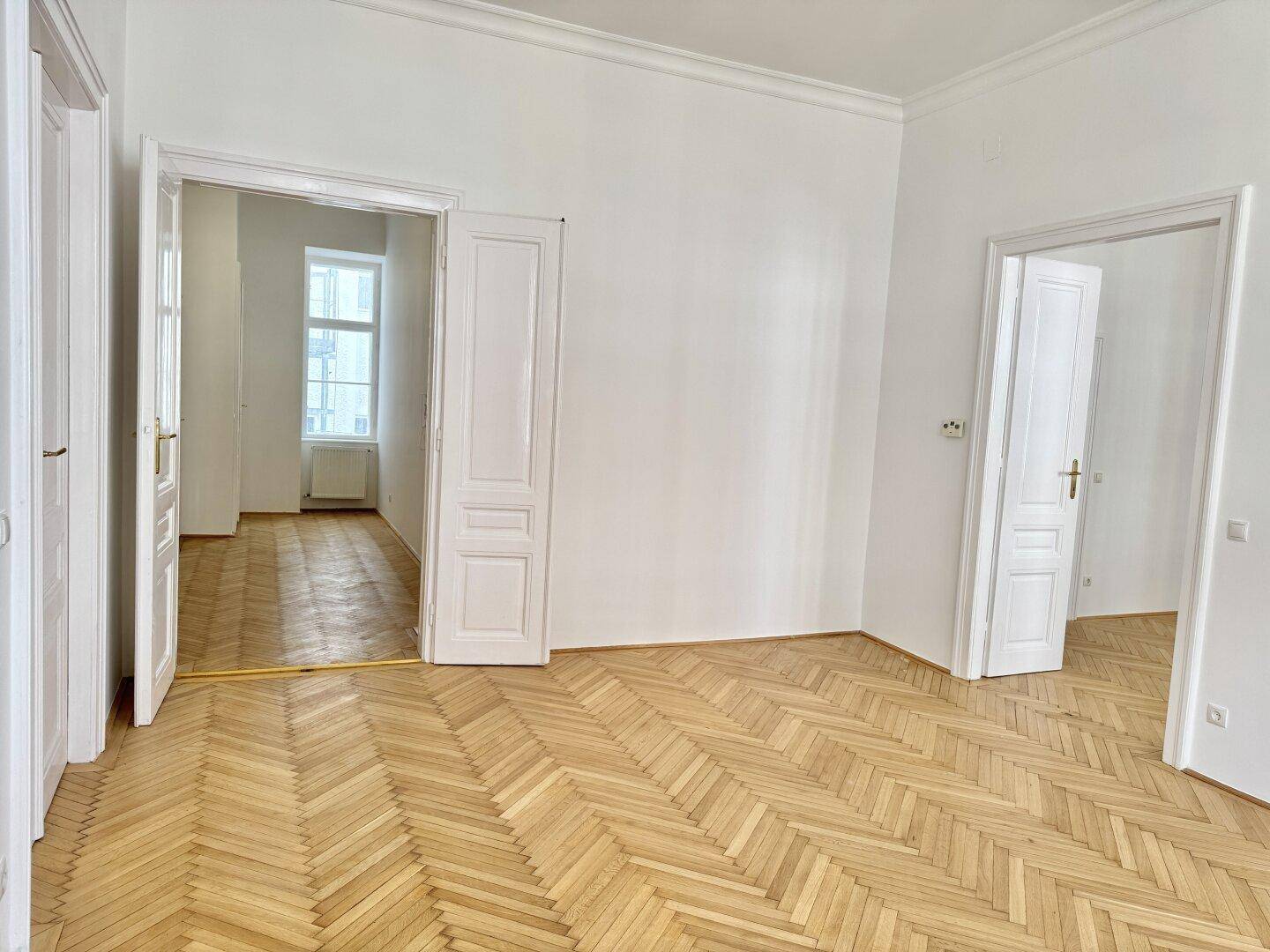 Großzügiger Raum mit Fischgrätparkett und hohen Decken, der durch offene Türen weitere Zimmer verbindet.