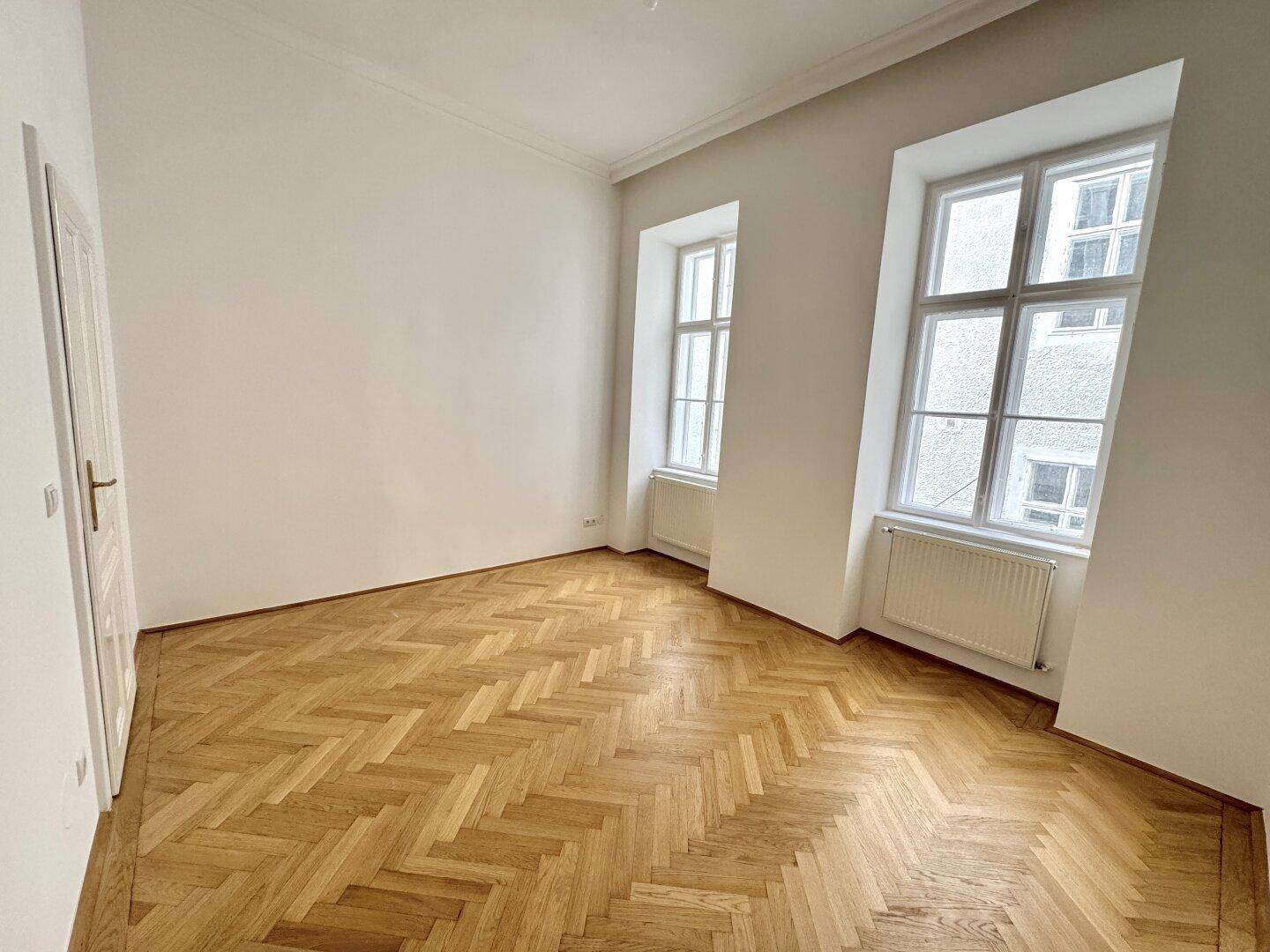 Geräumiges Zimmer mit Fischgrätparkett und zwei Fenstern, das viel Tageslicht in den Raum bringt.