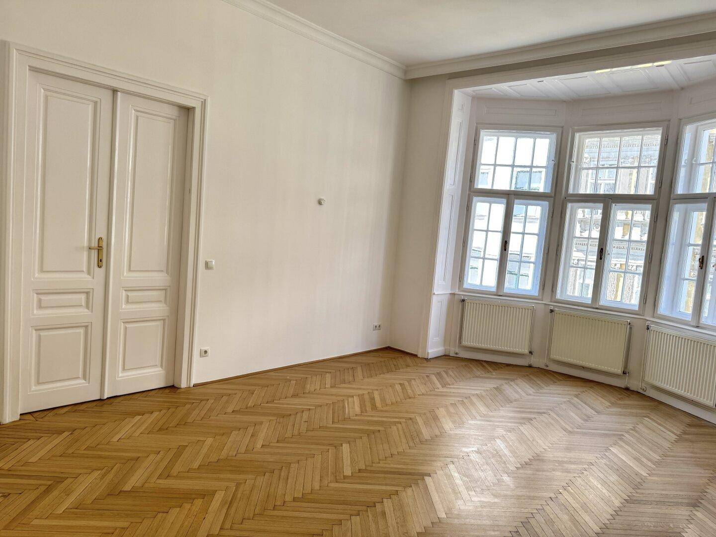 Geräumiges Zimmer mit Fischgrätparkett, hohen Decken und einem großen Erkerfenster, das viel Licht hereinlässt.