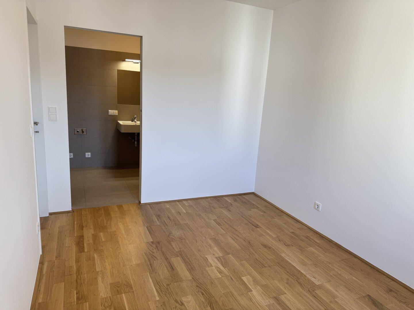 Heller Raum mit Holzfußboden und Blick in ein modern gefliestes Badezimmer mit Waschbecken.
