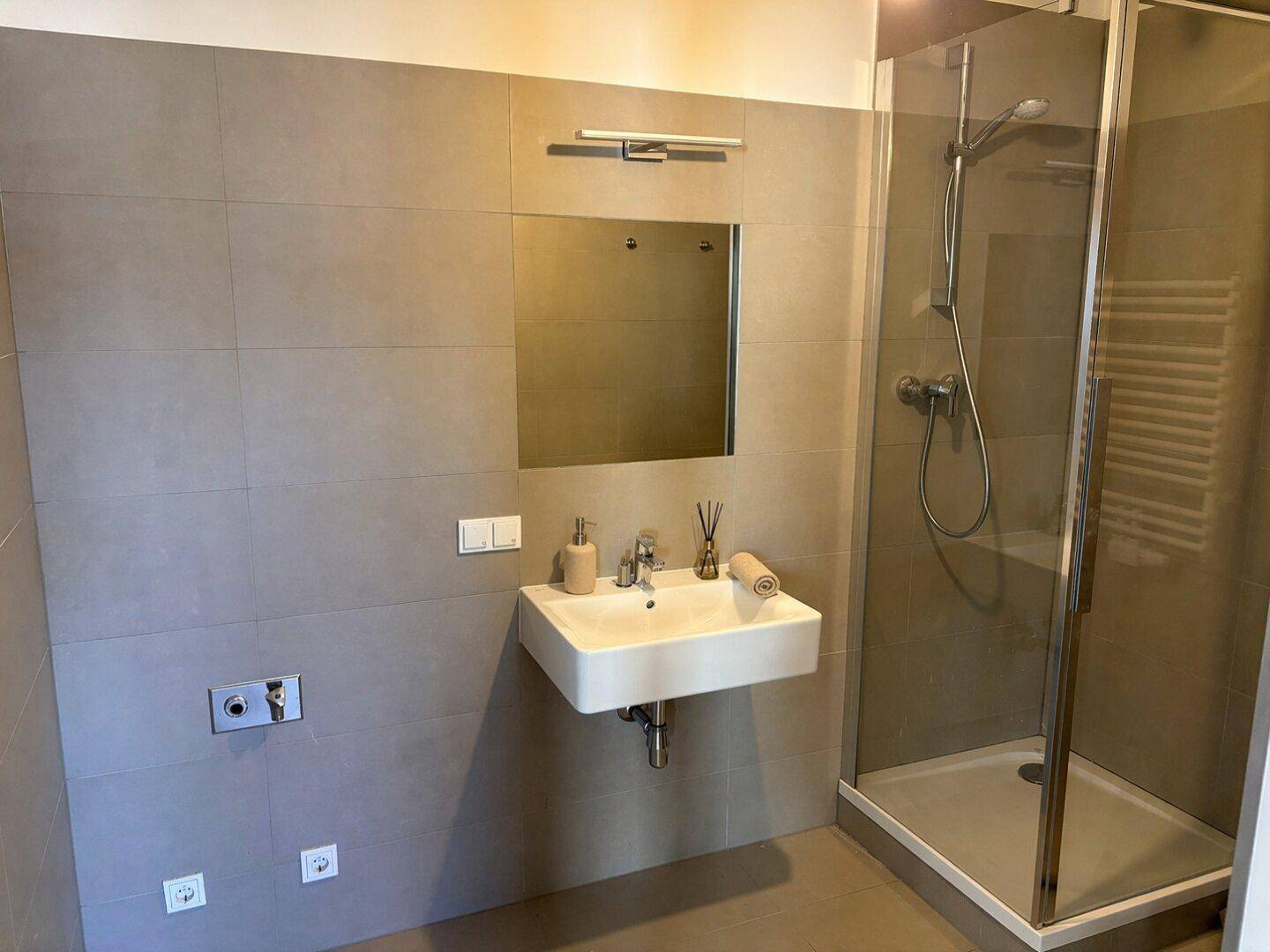 Modernes Badezimmer mit grauen Fliesen, wandhängendem Waschbecken, Spiegel und ebenerdiger Dusche.