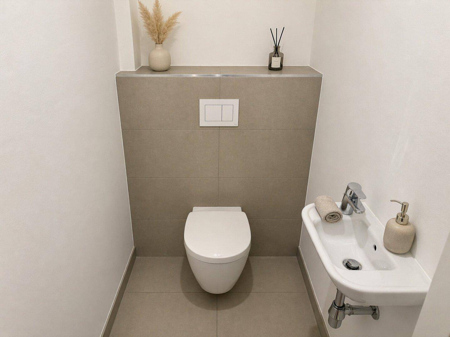 Modernes Gäste-WC mit wandhängender Toilette, kleinem Waschbecken und grauen Fliesen.