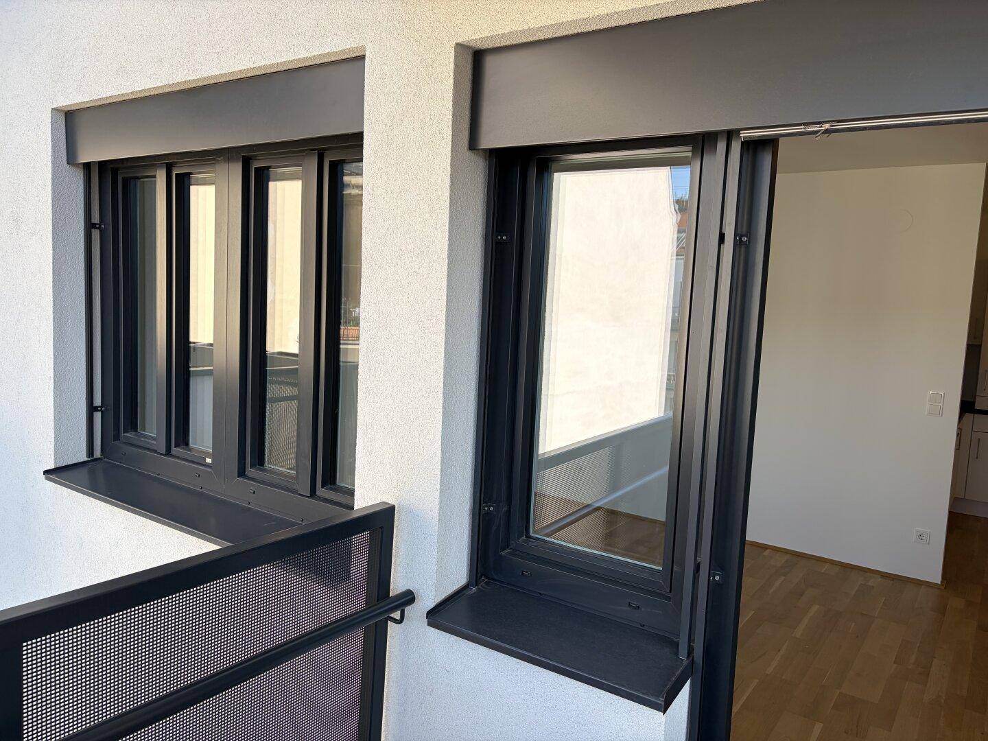 Moderner Balkon mit dunklen Fensterrahmen und Blick in den hellen Innenraum mit Parkettboden.