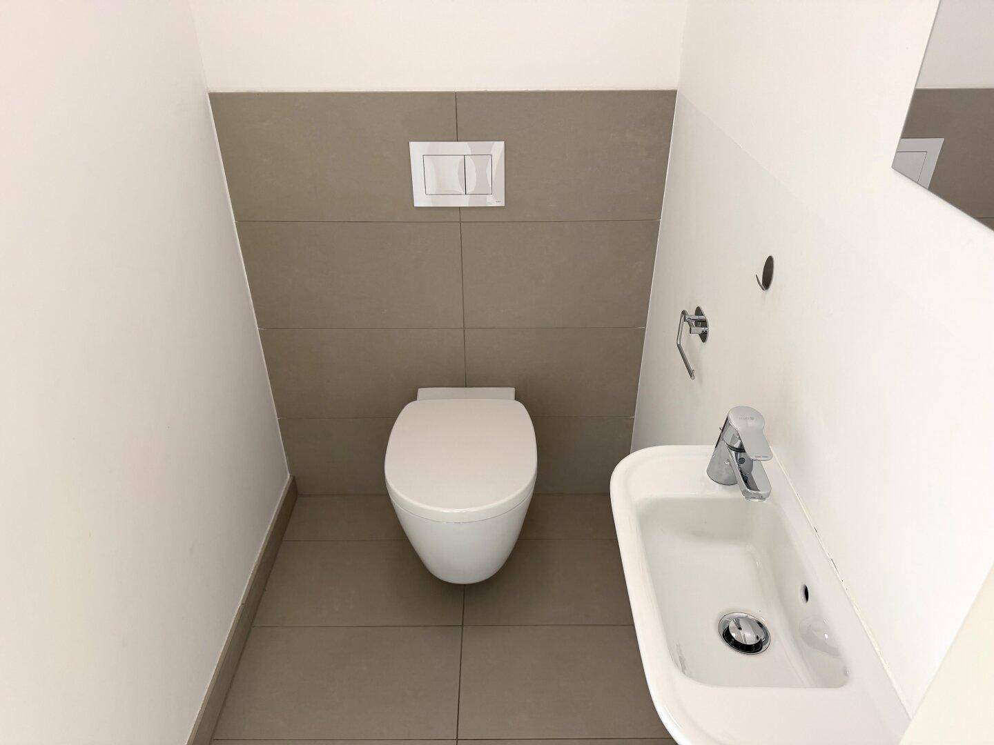 Separates WC mit wandhängender Toilette, kleinem Waschbecken und modernen Fliesen.