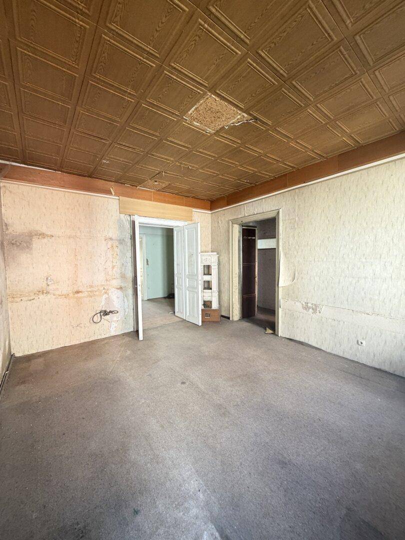 Leeres Zimmer mit alter Deckenverkleidung und abgenutztem Teppichboden.