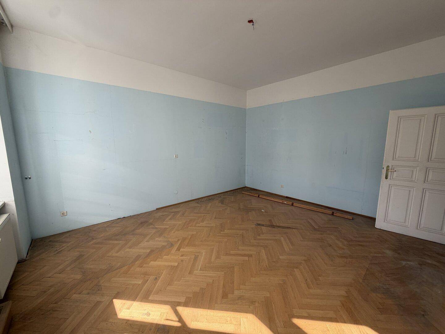Geräumiges Zimmer mit Fischgrätparkett und blauen Wänden, bereit für Renovierung.