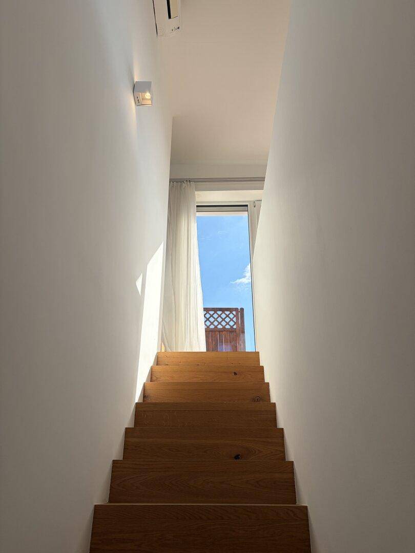 Helle Holztreppe führt zu einem oberen Bereich mit viel Tageslicht und Blick ins Freie.