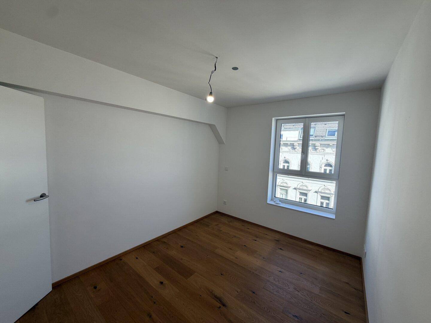Geräumiges Zimmer mit Holzboden und einem Fenster, das einen Blick auf die umliegenden Gebäude bietet.
