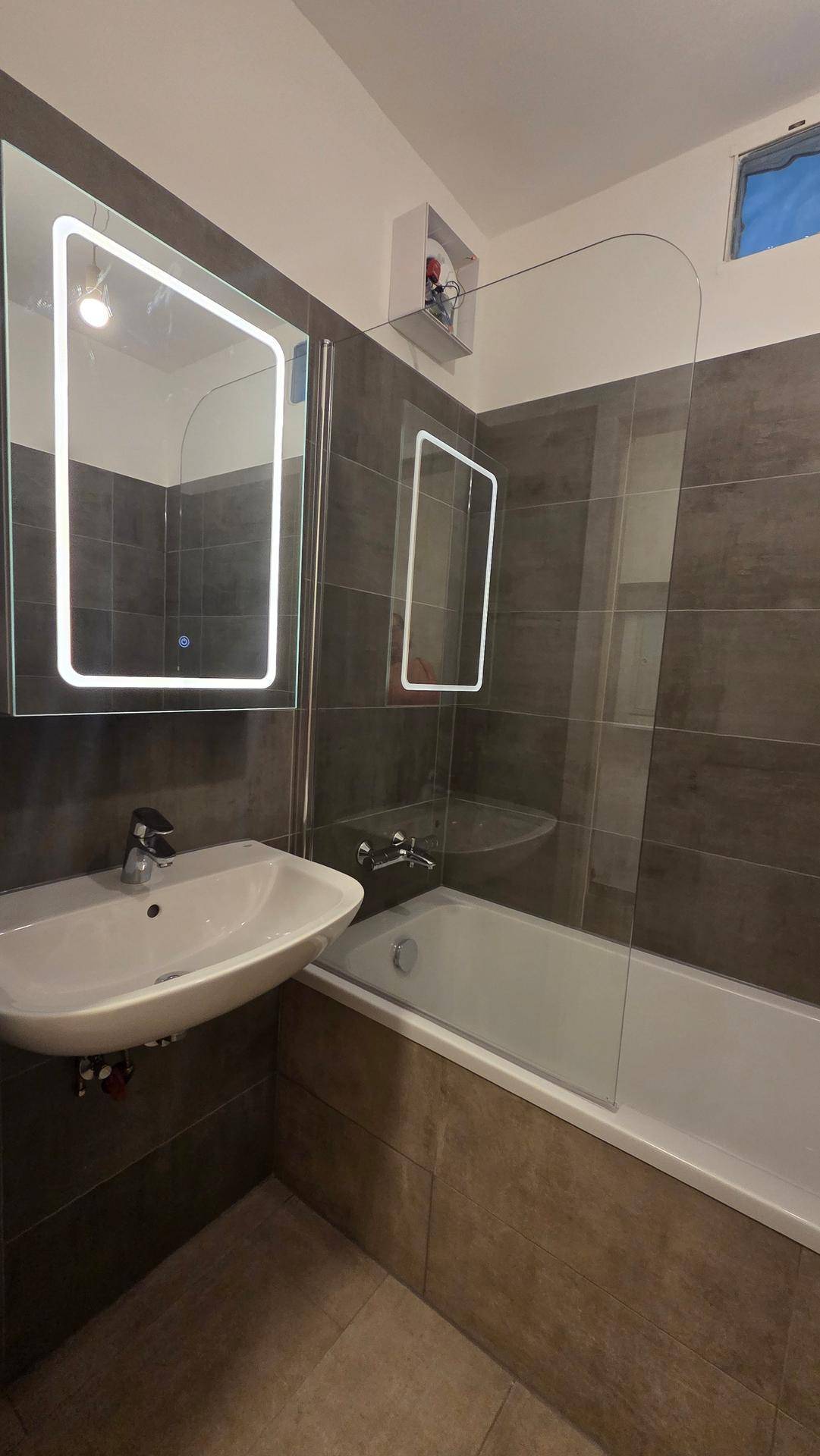 Modernes Badezimmer mit Badewanne, Duschabtrennung aus Glas, beleuchtetem Spiegel und kleinem Fenster.