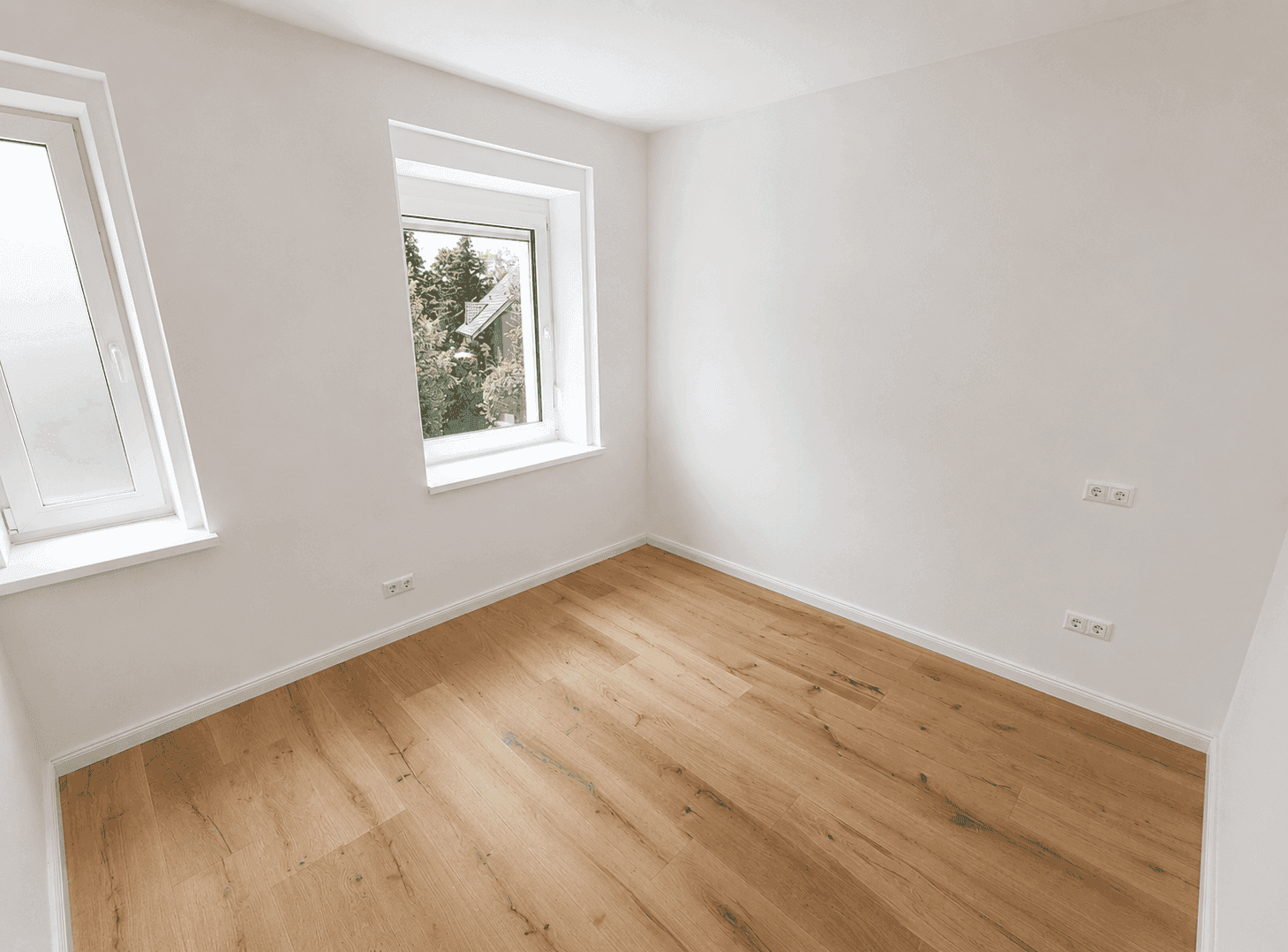Leerer Raum mit Holzboden und zwei Fenstern, die viel Tageslicht und einen grünen Ausblick bieten.