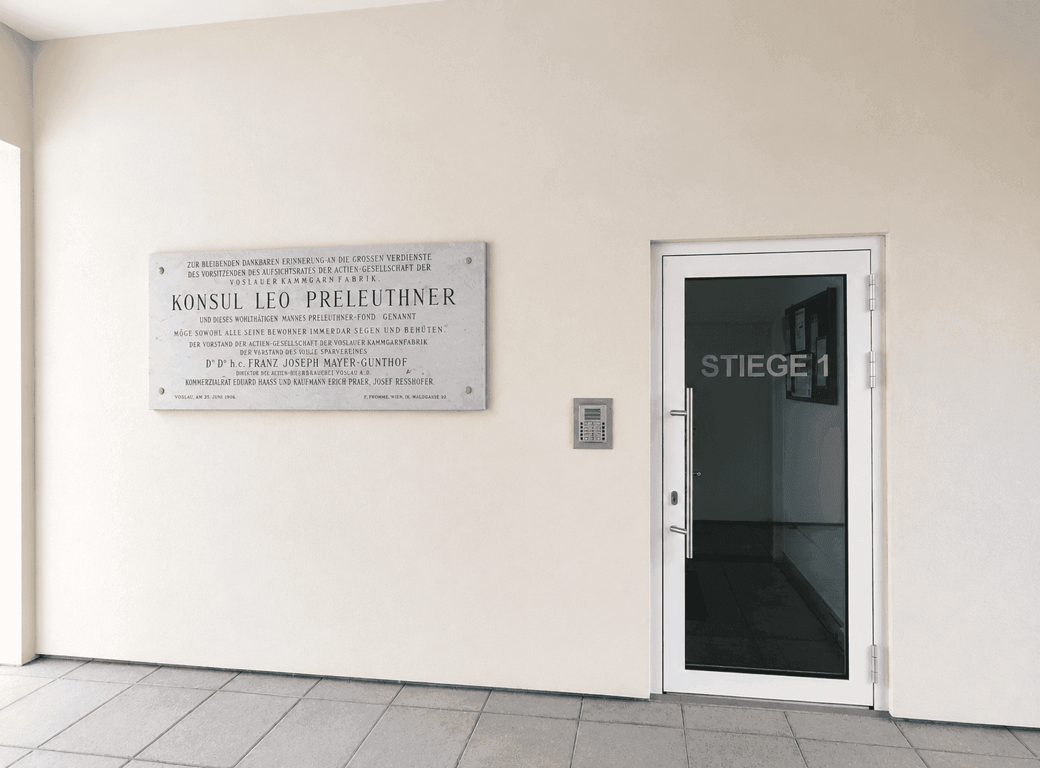 Gebäudeeingang mit Gedenktafel, moderner Tür und Gegensprechanlage.