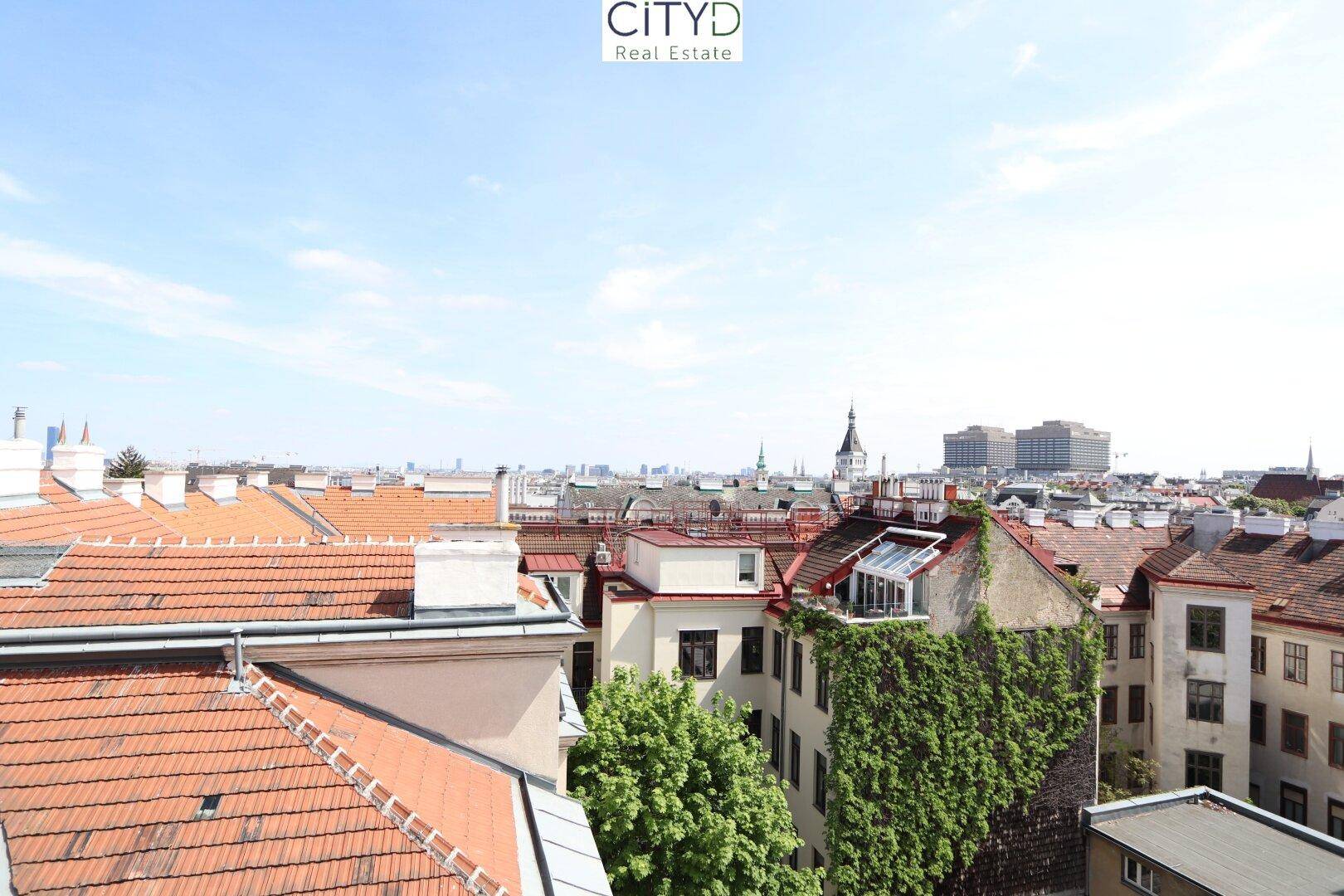 Panoramablick über die Dächer der Stadt mit grünen Bäumen und historischen Gebäuden unter blauem Himmel.